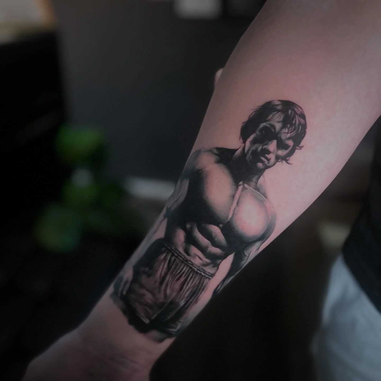 Black and gray realistic tattoo of a young male boxer with gloves, shorts, and an intense expression on his face, on a person's forearm.