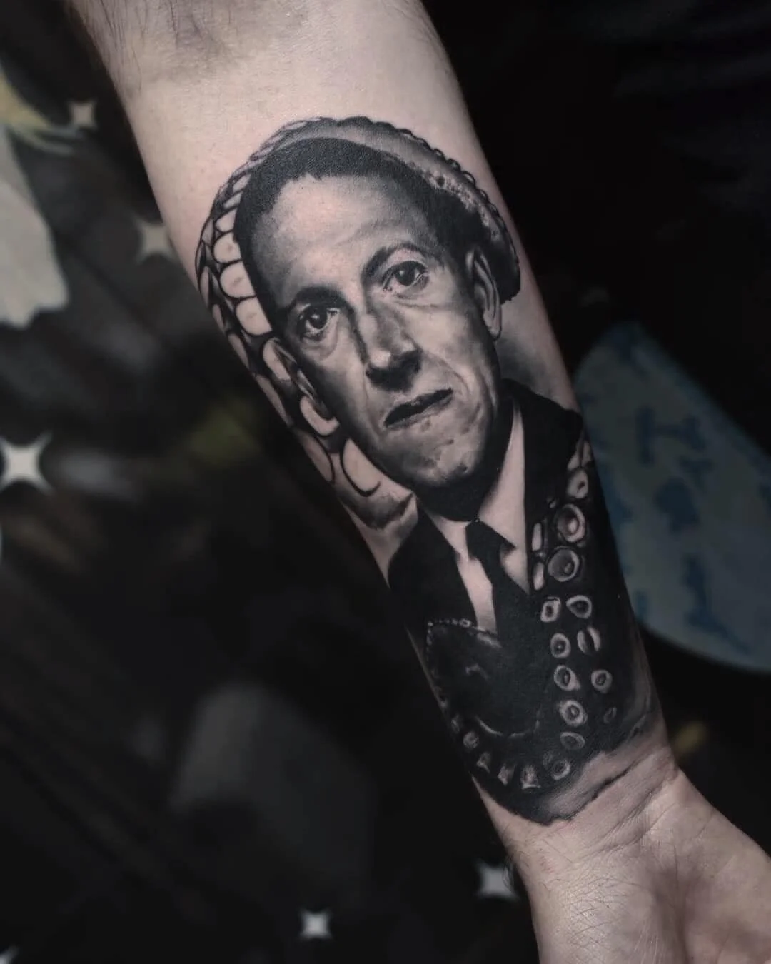 Black and white tattoo of a man with a serious expression, slicked-back hair, wearing a suit and tie, with a large octopus wrapping around his head and shoulders.