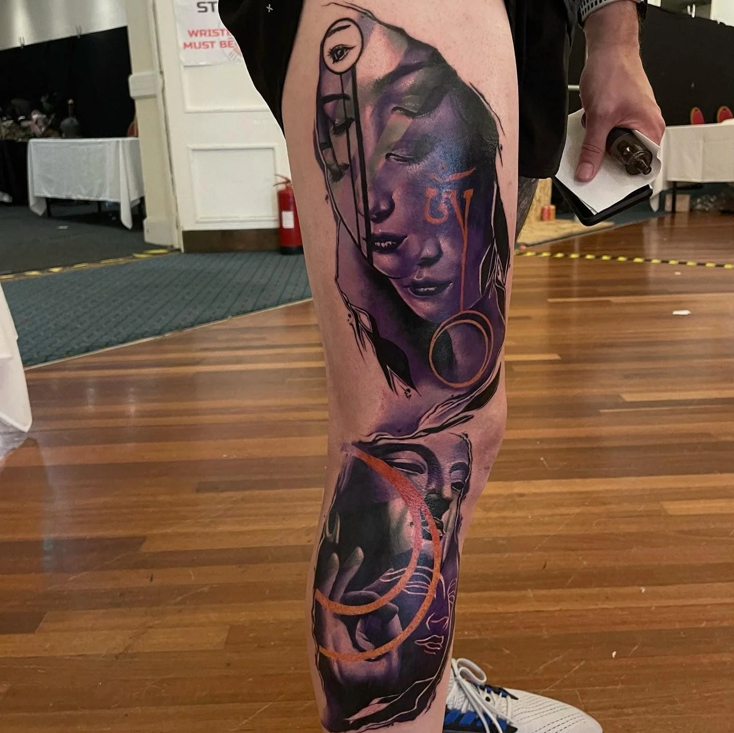 A person's leg with a colorful tattoo featuring multiple faces and abstract elements, including a woman with closed eyes and additional facial details, all rendered in purple, black, and orange ink.