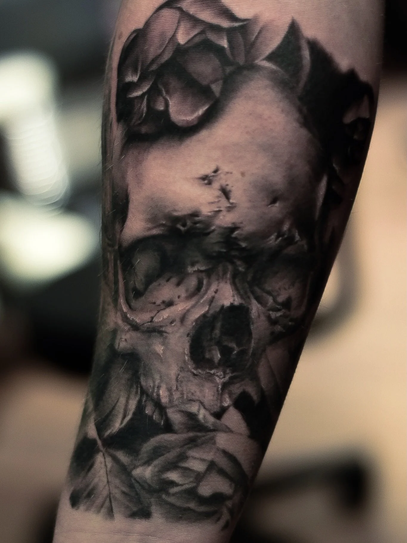 Black and gray tattoo of a realistic face with closed eyes, surrounded by roses and leaves, on a person's arm.