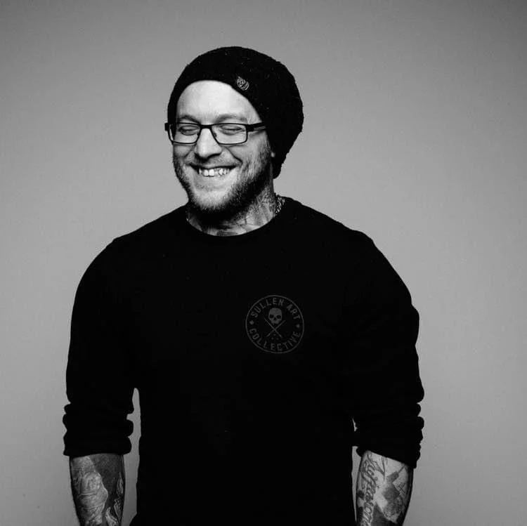 Black and white photo of a smiling man with glasses, a beanie hat, a dark long-sleeve shirt, and tattoos on his arms.