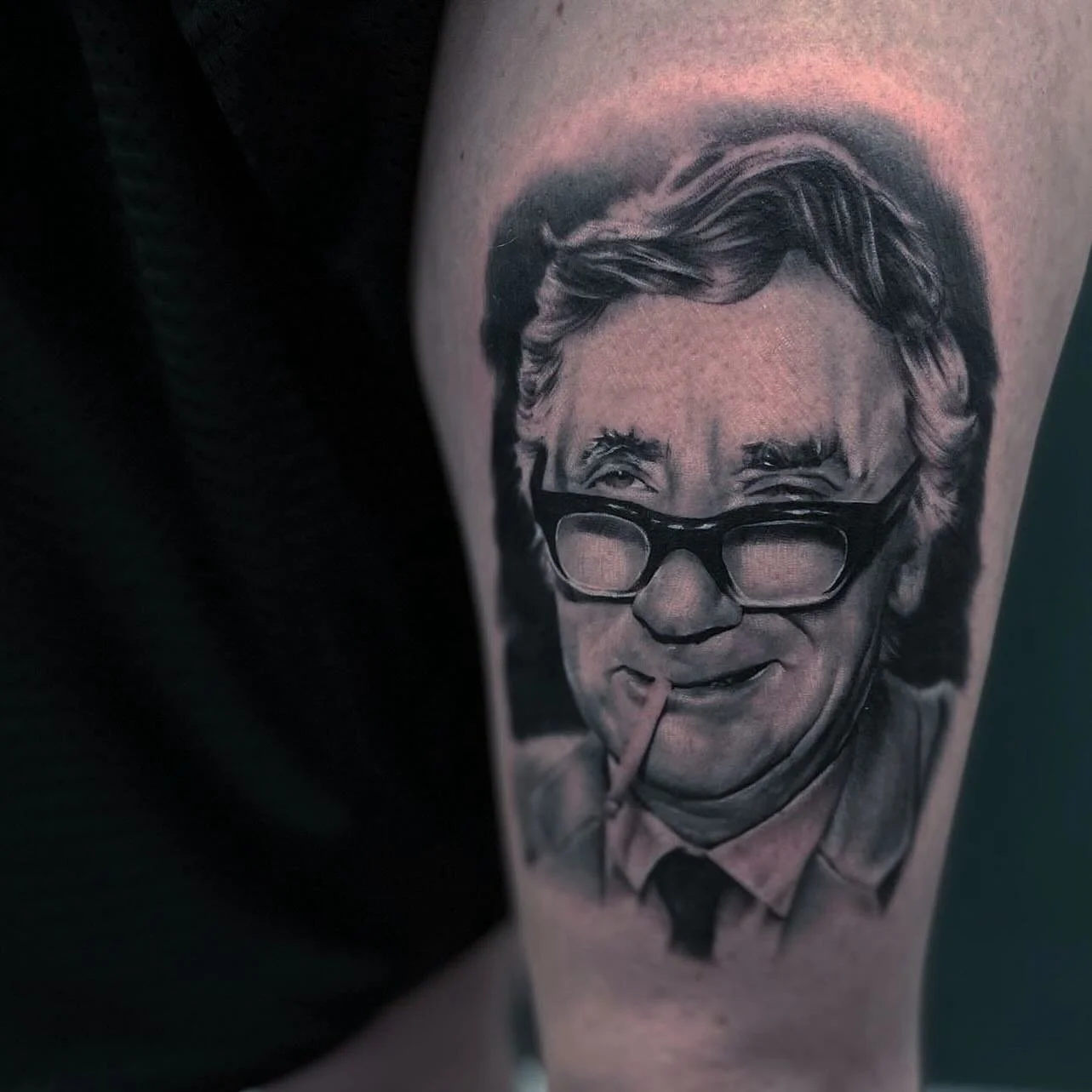 Black and gray tattoo of a man with glasses and a cigarette in his mouth, smiling.