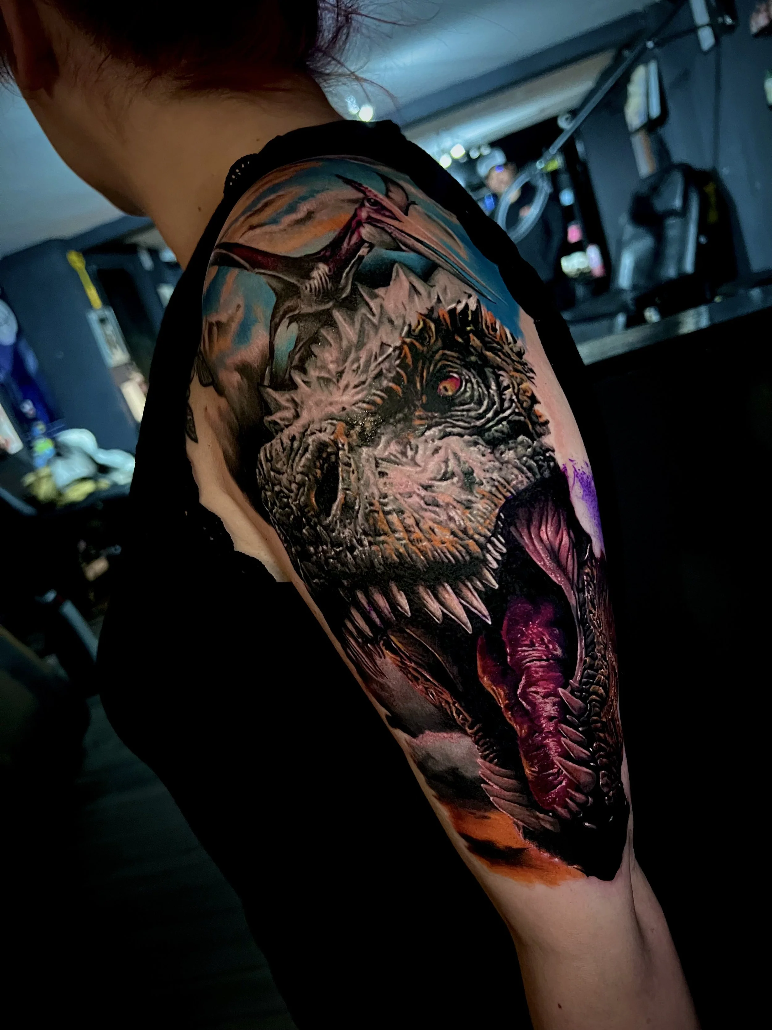 Colorful tattoo of a fierce dinosaur with open mouth, sharp teeth, and red eye on a person's upper arm and shoulder, with a background of clouds and sky.
