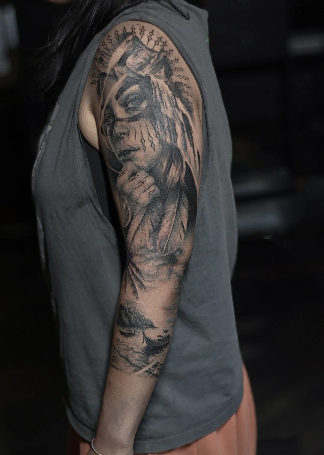 Black and gray tattoo sleeve featuring a woman's face with long hair, a mountain scene, ocean waves, a boat, and a hut.
