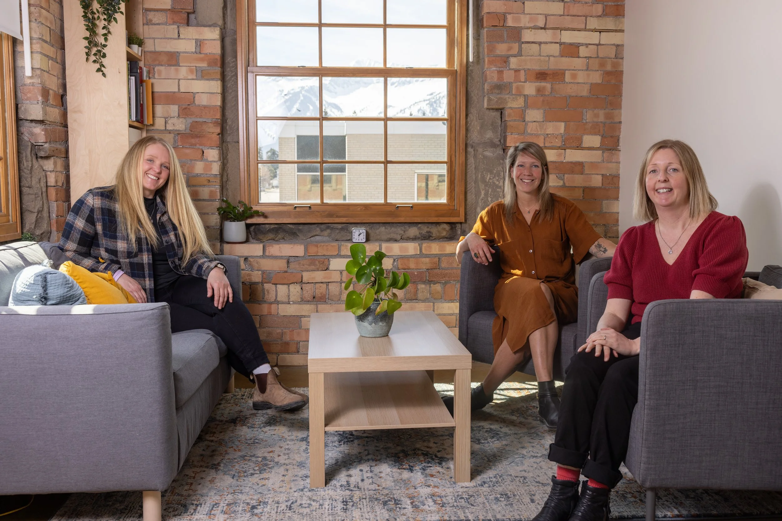 Three women sitting on gray sofas in a cozy room with exposed brick walls, a large window with mountain views, and a small coffee table with a potted plant.