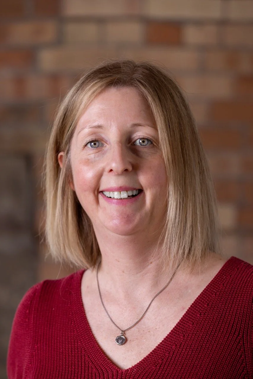A woman with blonde hair, blue eyes, and fair skin, smiling, wearing a red top and a silver necklace, in front of a brick wall background.