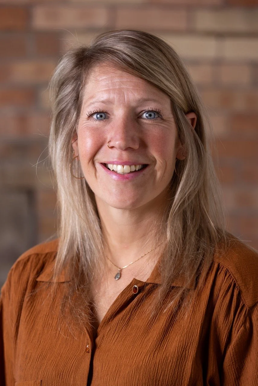 A woman with blonde hair, blue eyes, and a nose ring, smiling in front of a brick wall, wearing a brown blouse and a gold necklace.