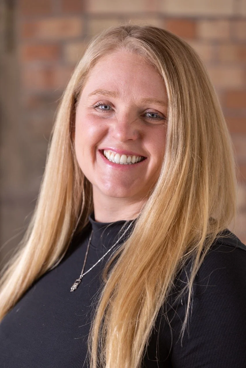 A smiling woman with long blonde hair, wearing a black turtleneck and a plaid shirt, in front of a brick wall.