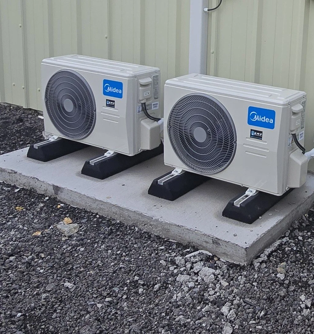 Two white Midea air conditioning outdoor units installed on a raised concrete pad outside a building with light green siding.