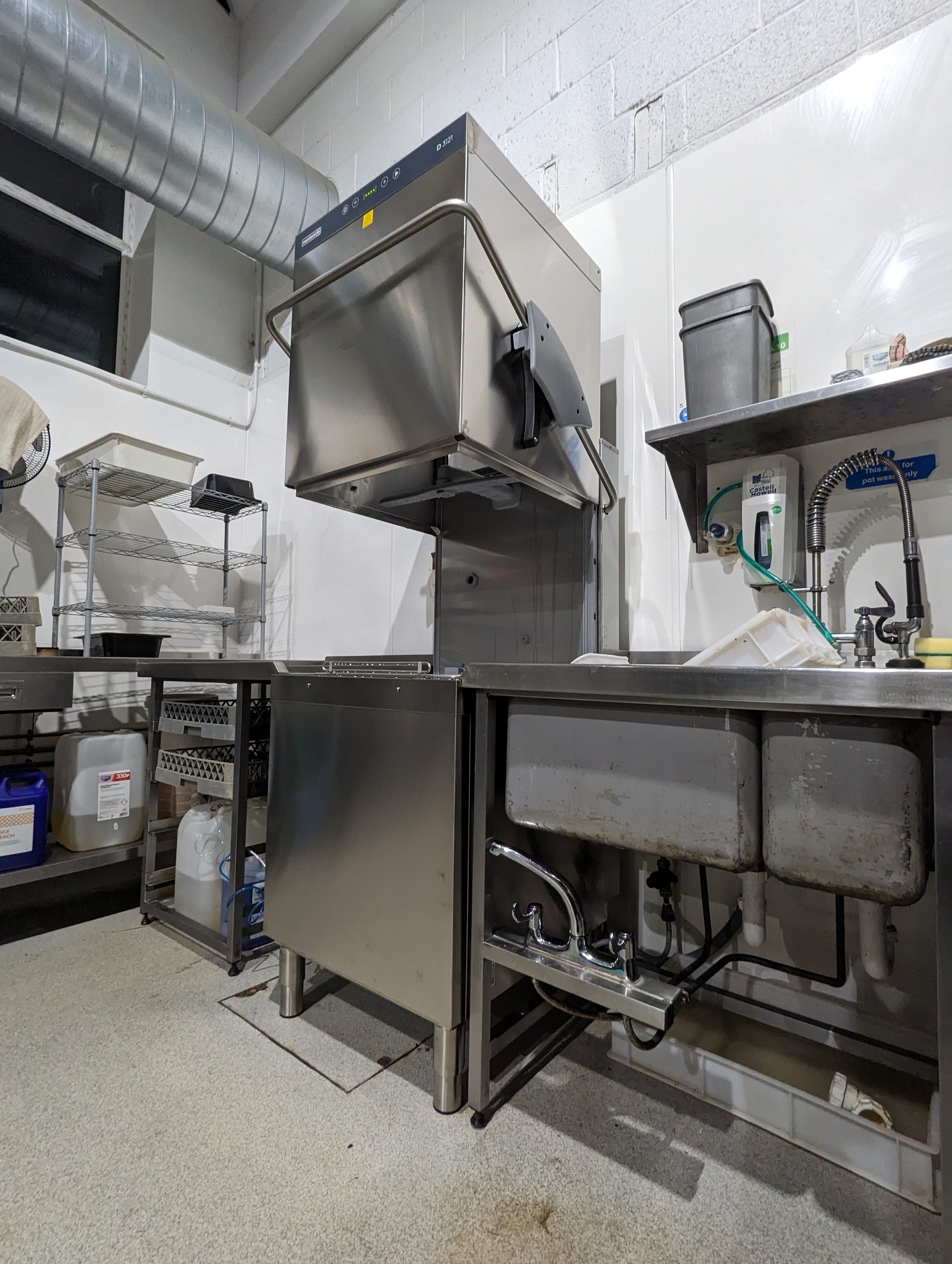 Industrial kitchen sink with a large stainless steel dishwasher and shelves with supplies, located in a commercial or industrial setting.
