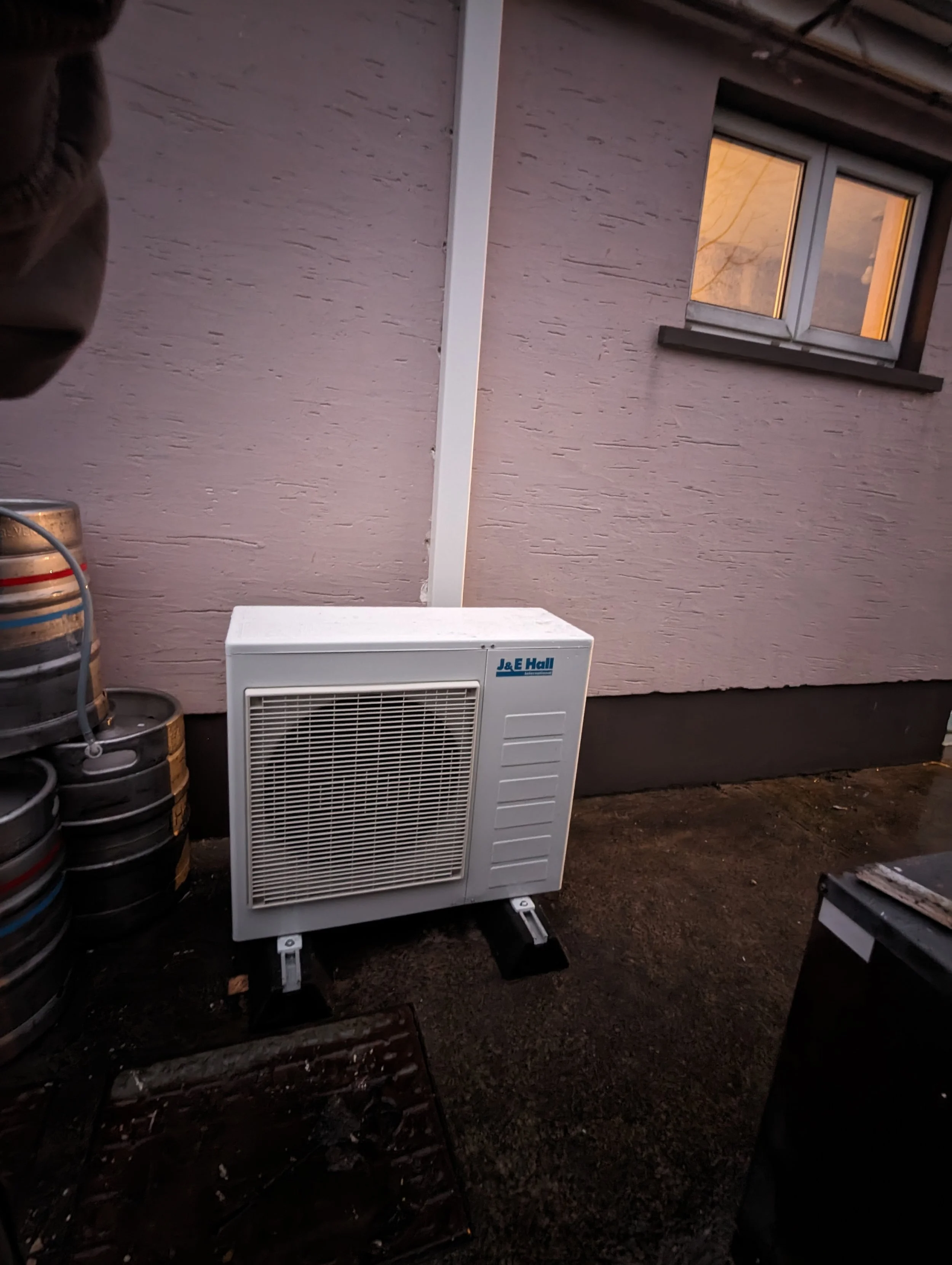 A white outdoor air conditioning unit on a metal stand outside a pink stucco house with a small window. There are several stacked drink kegs to the left and a black container at the bottom right corner.