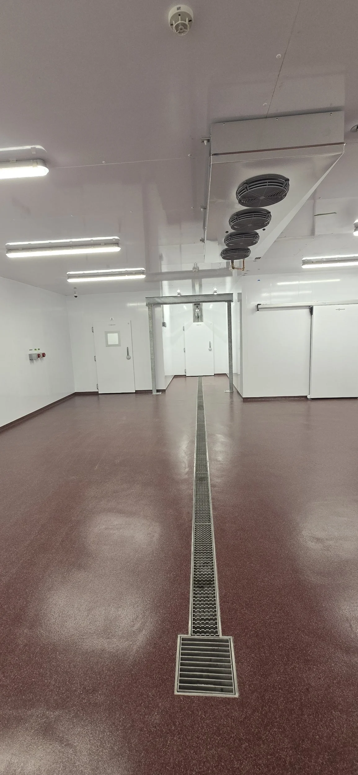 Clean industrial room with red textured flooring, white walls, ceiling-mounted ventilation fans, and a central drainage grate running down the middle of the floor.