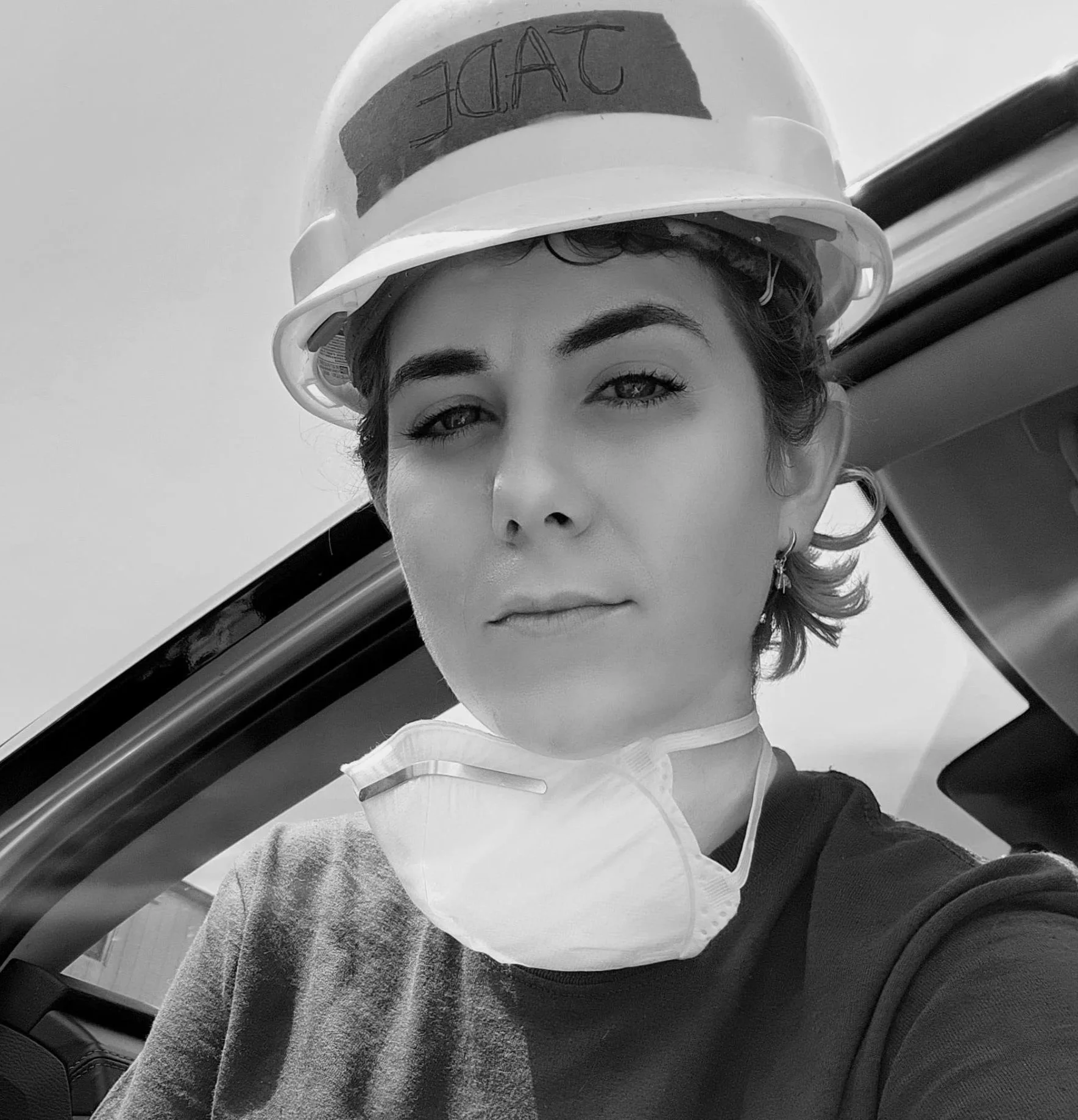 A woman wearing a hard hat with the word 'SAUCE' written on it, a face mask hanging under her chin, and earrings, sitting inside a vehicle.