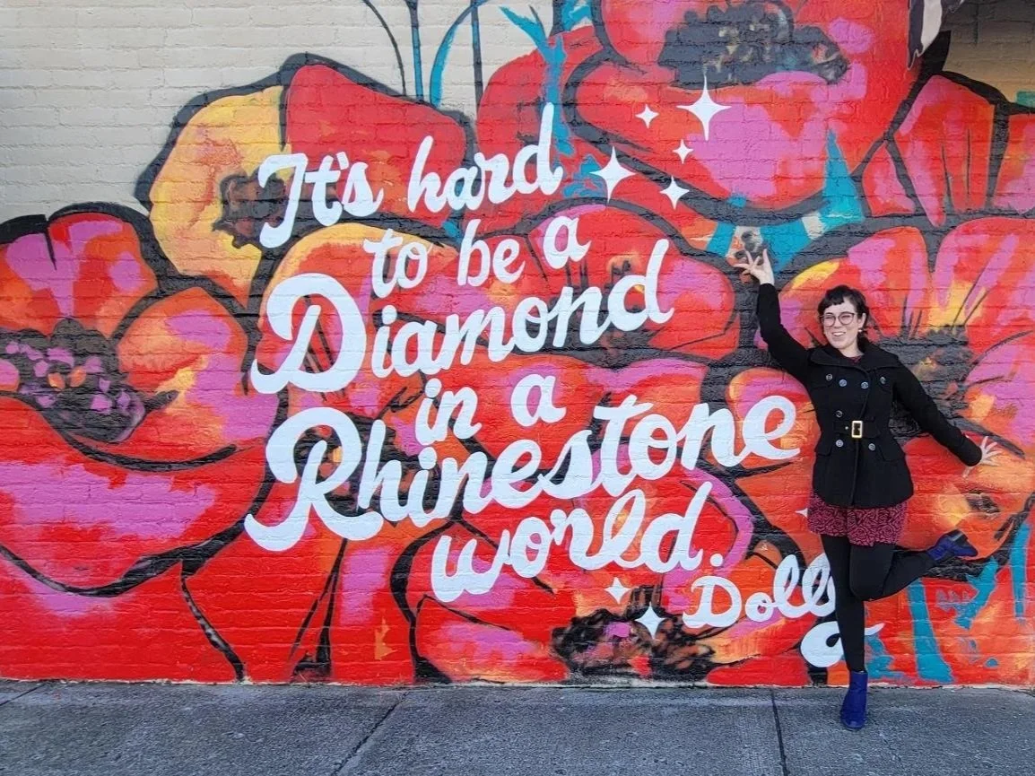 A woman with glasses and a black coat standing in front of a colorful graffiti wall with large red, pink, yellow, and purple flowers and the text 'It's hard to be a diamond in a rhinestone world' written in white cursive letters.