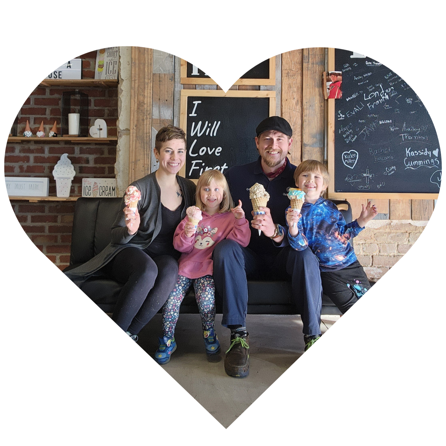 A family of five sitting on a black couch in an ice cream shop, holding ice cream cones and smiling at the camera. The background has chalkboards with writing and a brick wall with shelves decorated with ice cream-themed décor.