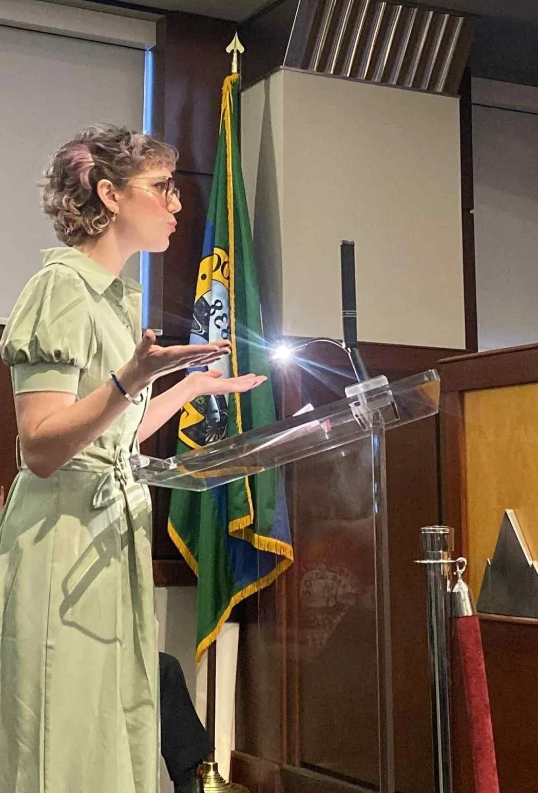 A woman with short curly hair, glasses, and earrings standing at a clear podium, gesturing with her hands. Behind her are blue, green, and yellow flags and a wood-paneled wall.