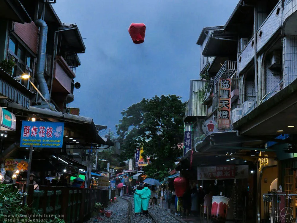Street scene with people walking with umbrellas, narrow street flanked by shops, colorful signs, trees in the background, and a sky lantern floating overhead.