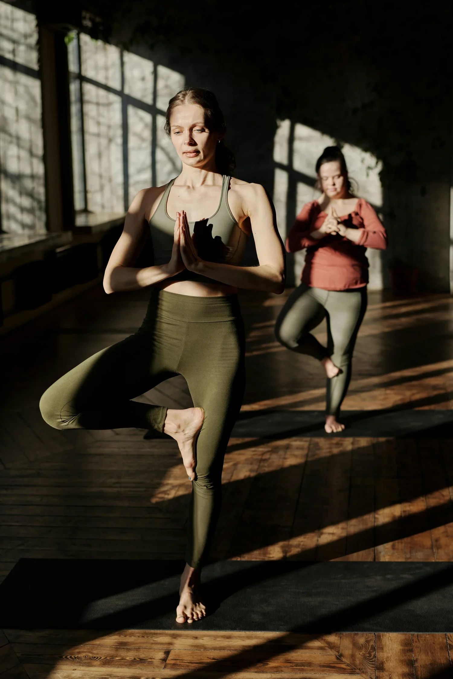 Two women practicing yoga indoors, standing on one leg with hands in a prayer position, with sunlight streaming through large windows, casting shadows on the wooden floor.