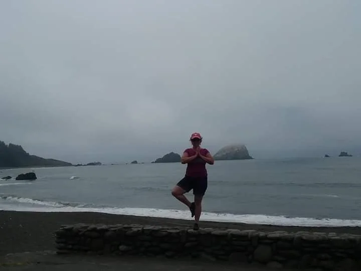 A person practicing yoga on a stone wall at a beach under an overcast sky with rocky islands in the background.