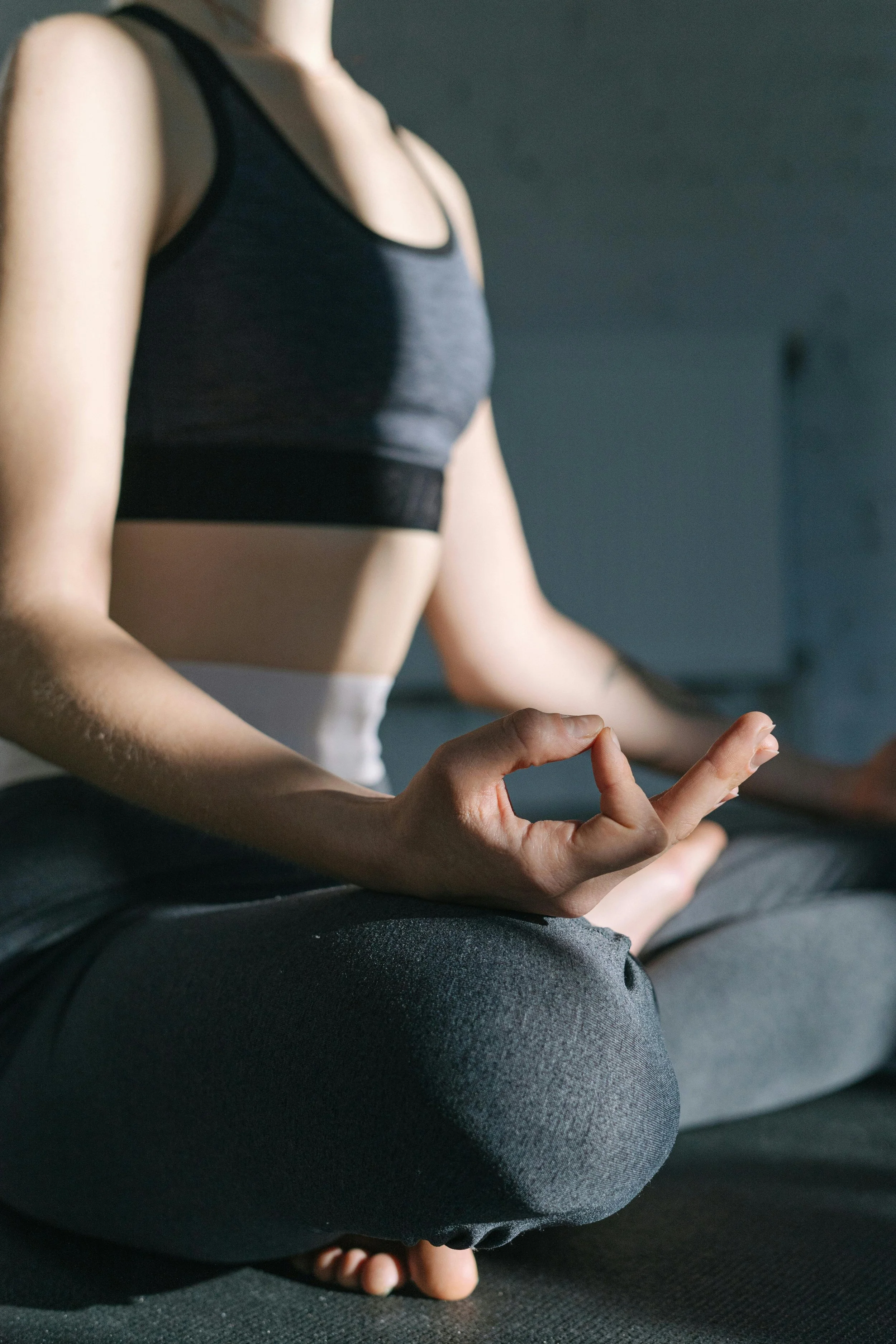 Close-up of a person sitting cross-legged on a yoga mat, wearing a black sports bra and dark leggings, practicing meditation with hands resting on knees and fingers in Gyan mudra. The background is plain and dark.