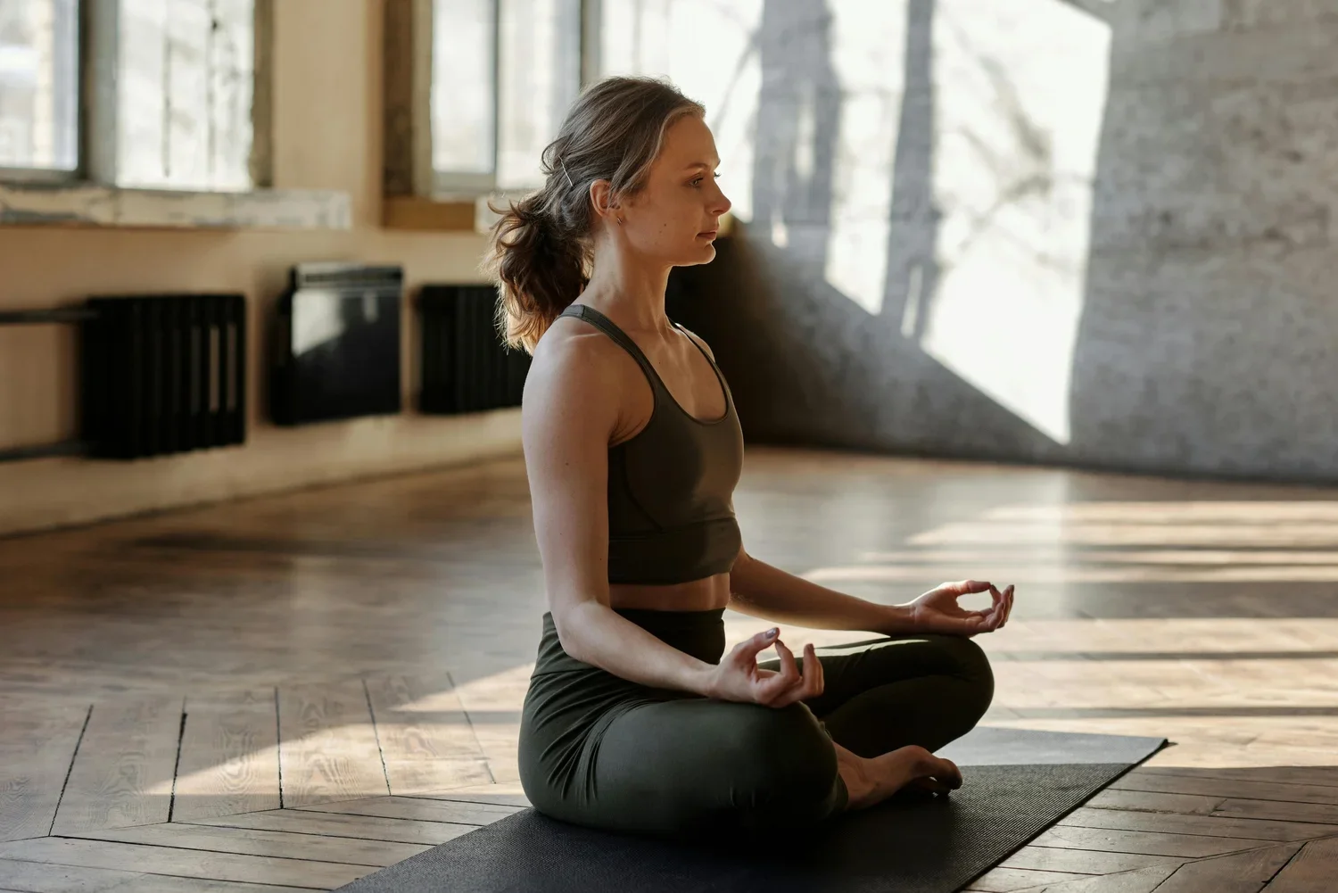 A woman practicing meditation in a bright, spacious room with wooden floors and large windows, sitting cross-legged on a yoga mat with hands in a mudra position.