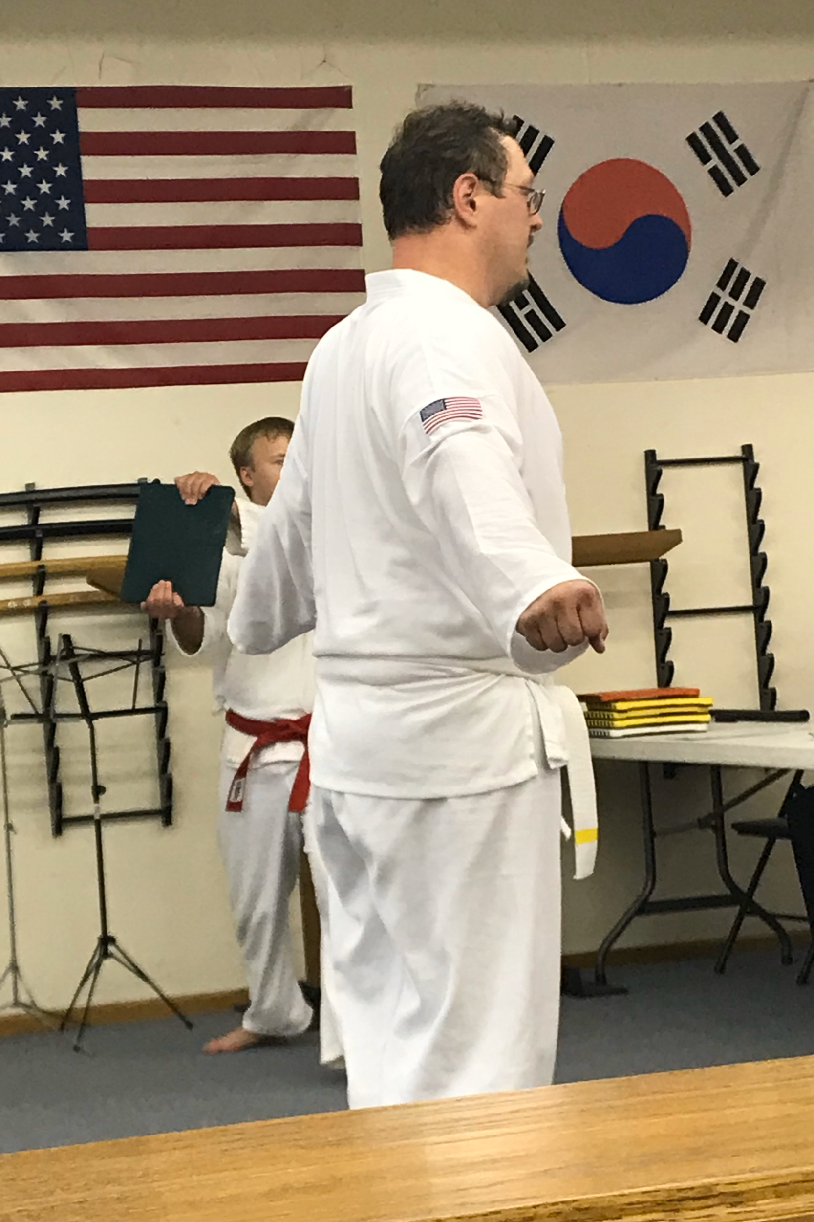 A man in a white martial arts uniform with an American flag patch on his arm demonstrating an attention stance in a room with American and South Korean flags