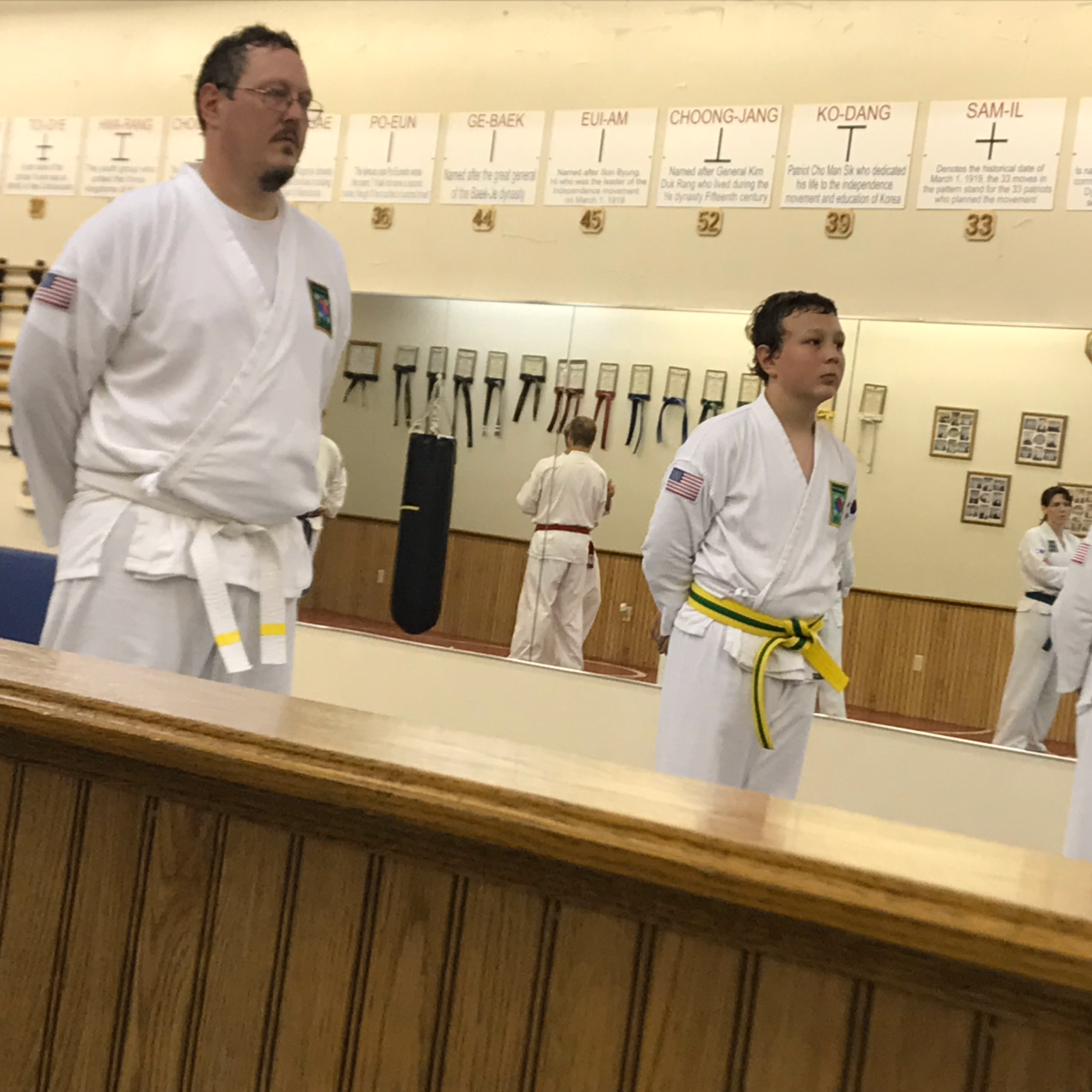 Martial arts students practicing in a dojo, with two individuals in the foreground wearing white gi uniforms, one with a black belt and the other with a yellow belt. Posters and belts displayed on the wall in the background.