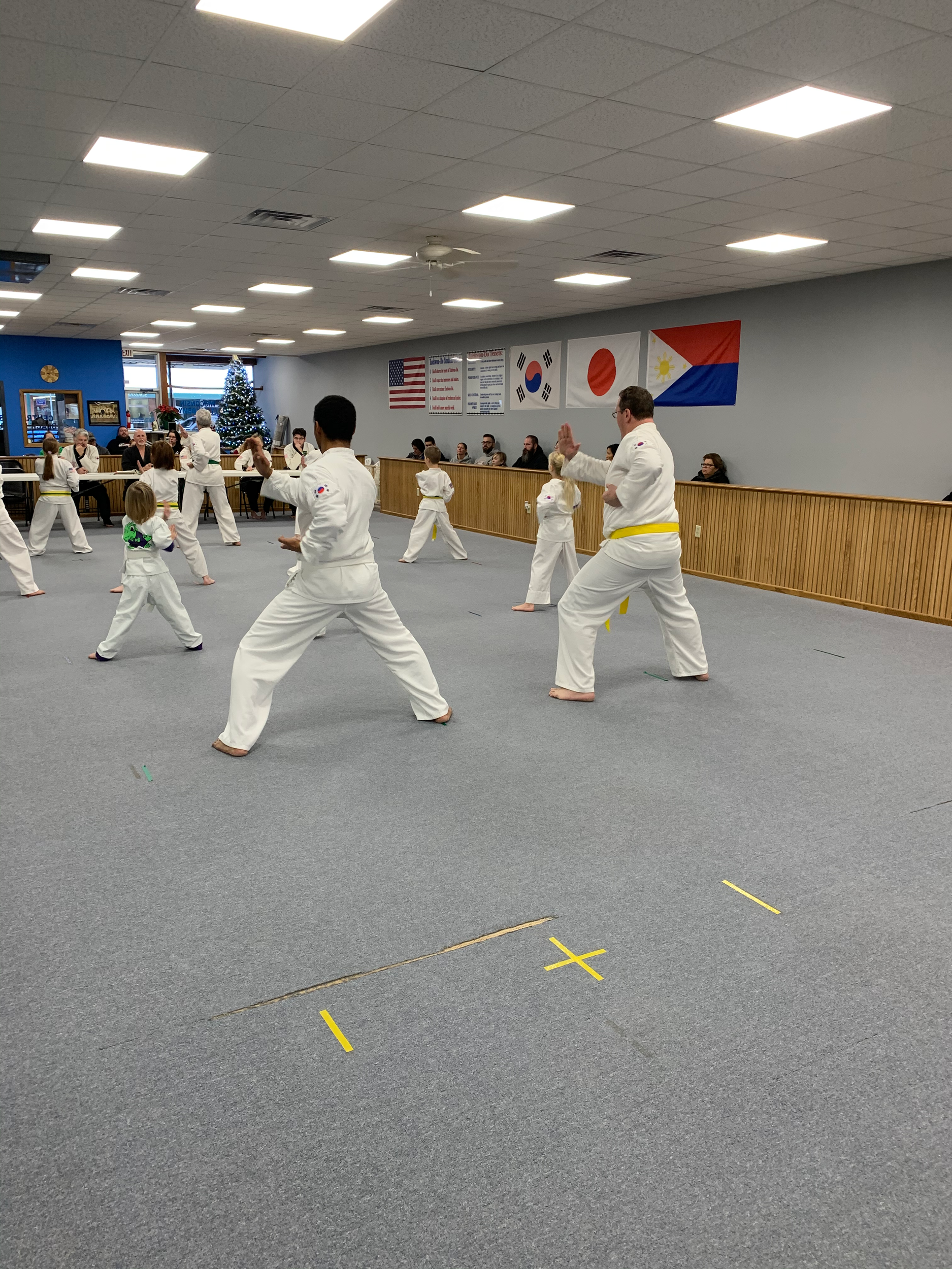 Martial arts class practicing in a dojang with flags of the United States, South Korea, Japan, and the Philippines on the wall, and a decorated Christmas tree in the background.