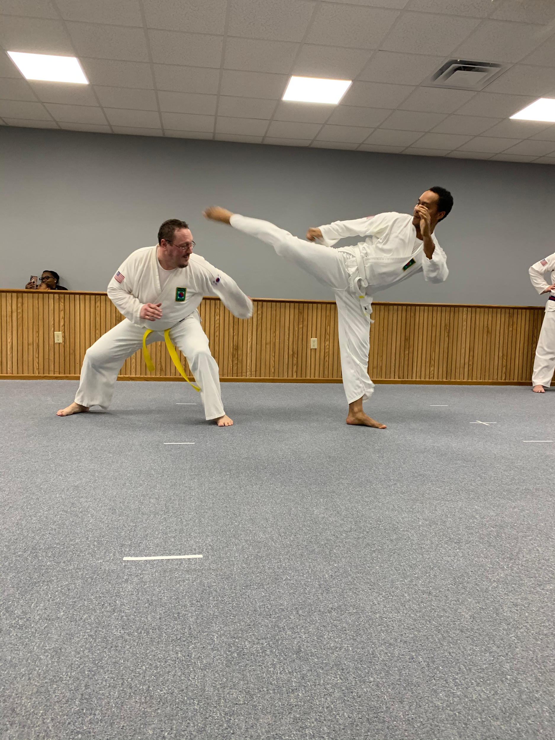 Two martial artists practicing in a dojo, one executing a high kick while the other prepares to block, both wearing traditional white gi uniforms with patches.