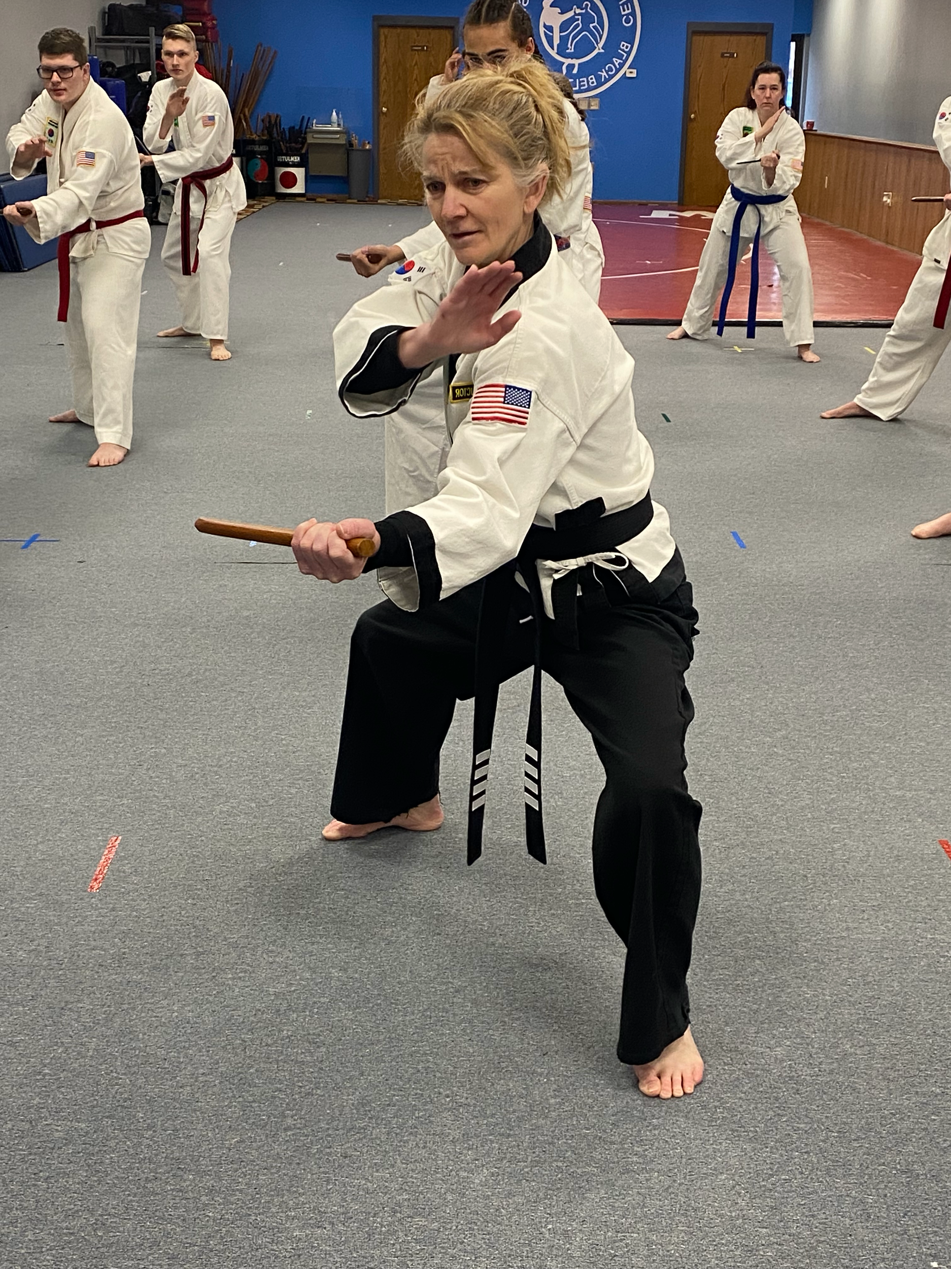 A woman in a martial arts uniform demonstrating a self-defense move in a martial arts training facility with children practicing in the background.