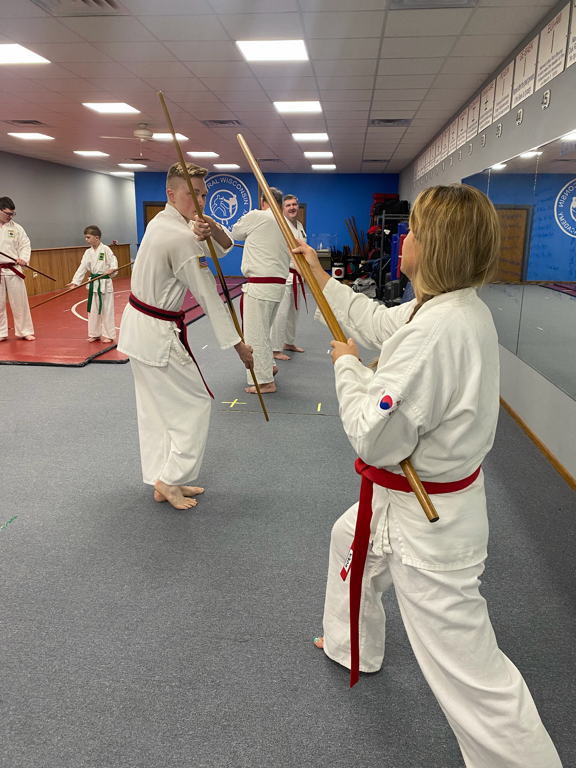 Martial arts students practicing in a dojo, wearing traditional uniforms with red belts, some holding wooden staffs.