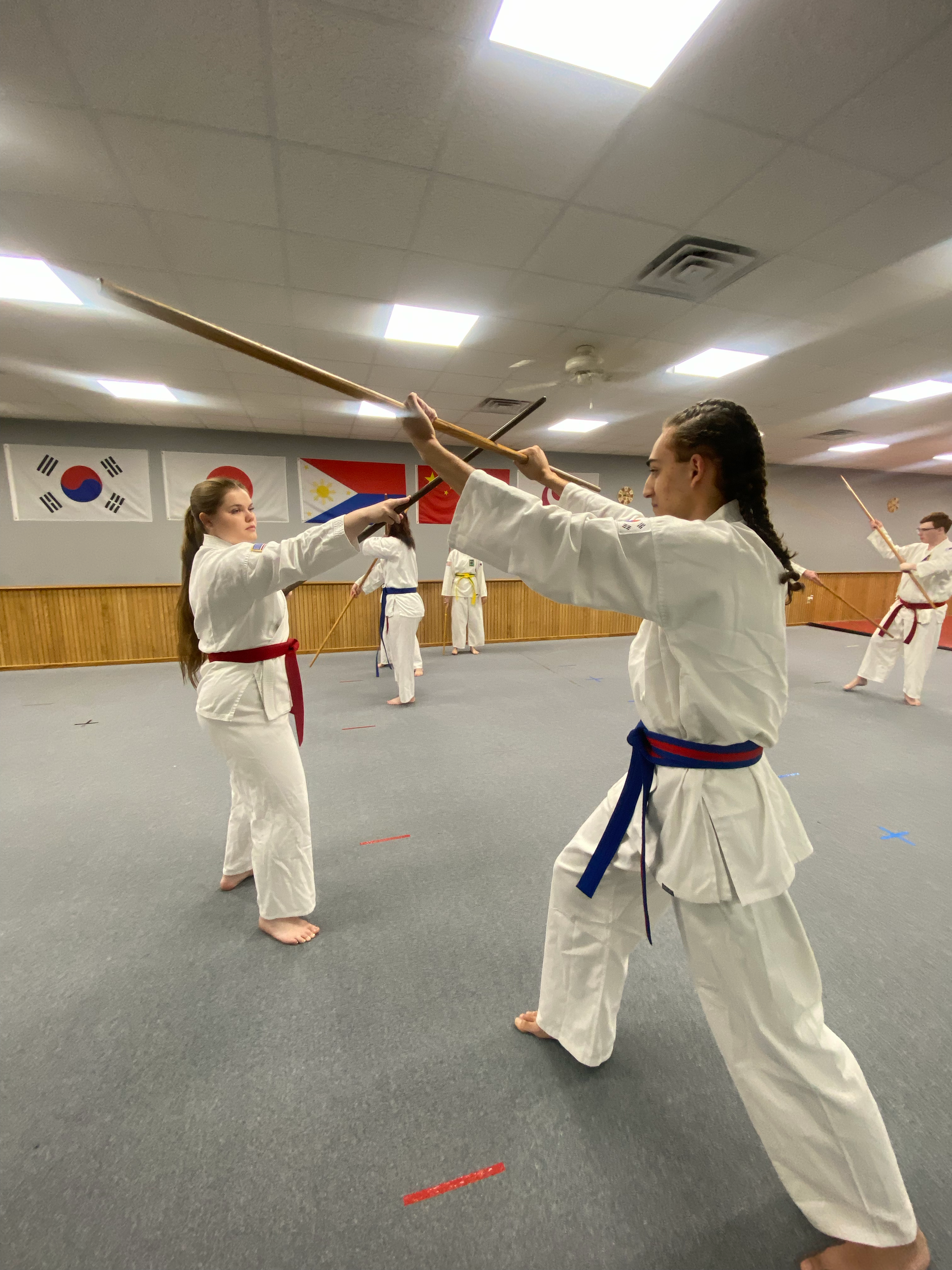 Two girls practicing martial arts in a dojo, holding wooden sticks and wearing traditional martial arts uniforms with colored belts. Other students are practicing in the background. Flags are displayed on the wall.