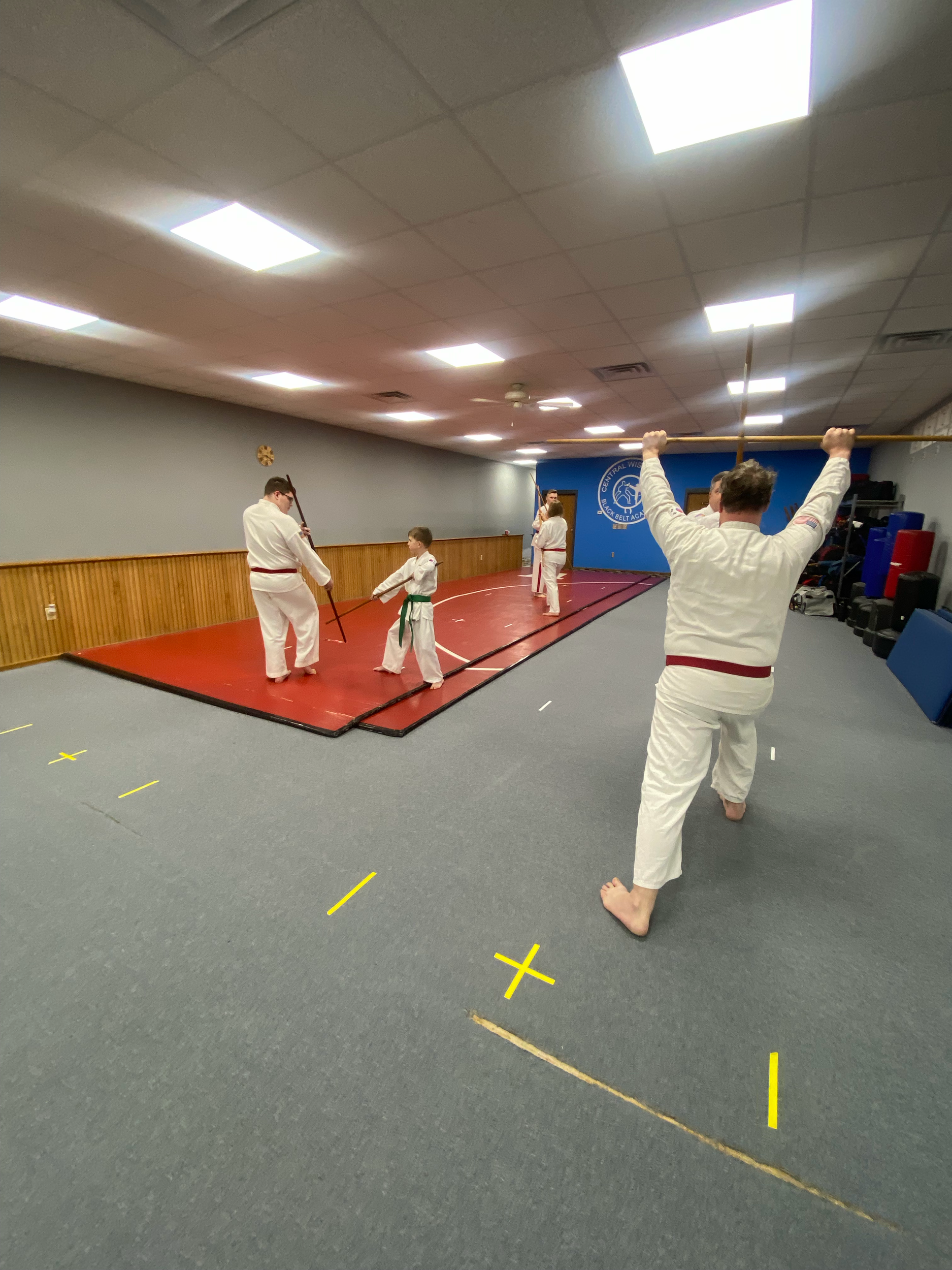 Martial arts class with children practicing with sticks on a red mat in a gym, supervised by an instructor in a white gi with a red belt.