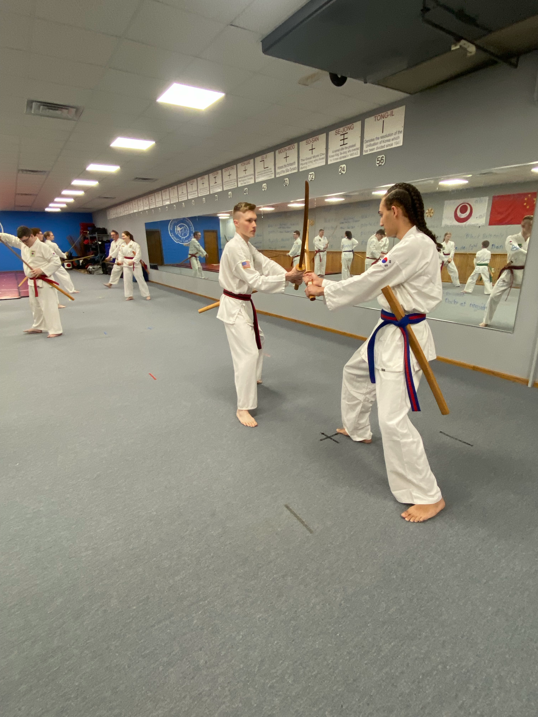 Two children practicing taekwondo in a martial arts dojo, wearing white uniforms with colored belts, and holding wooden weapons. Other children in uniforms are visible in the background.