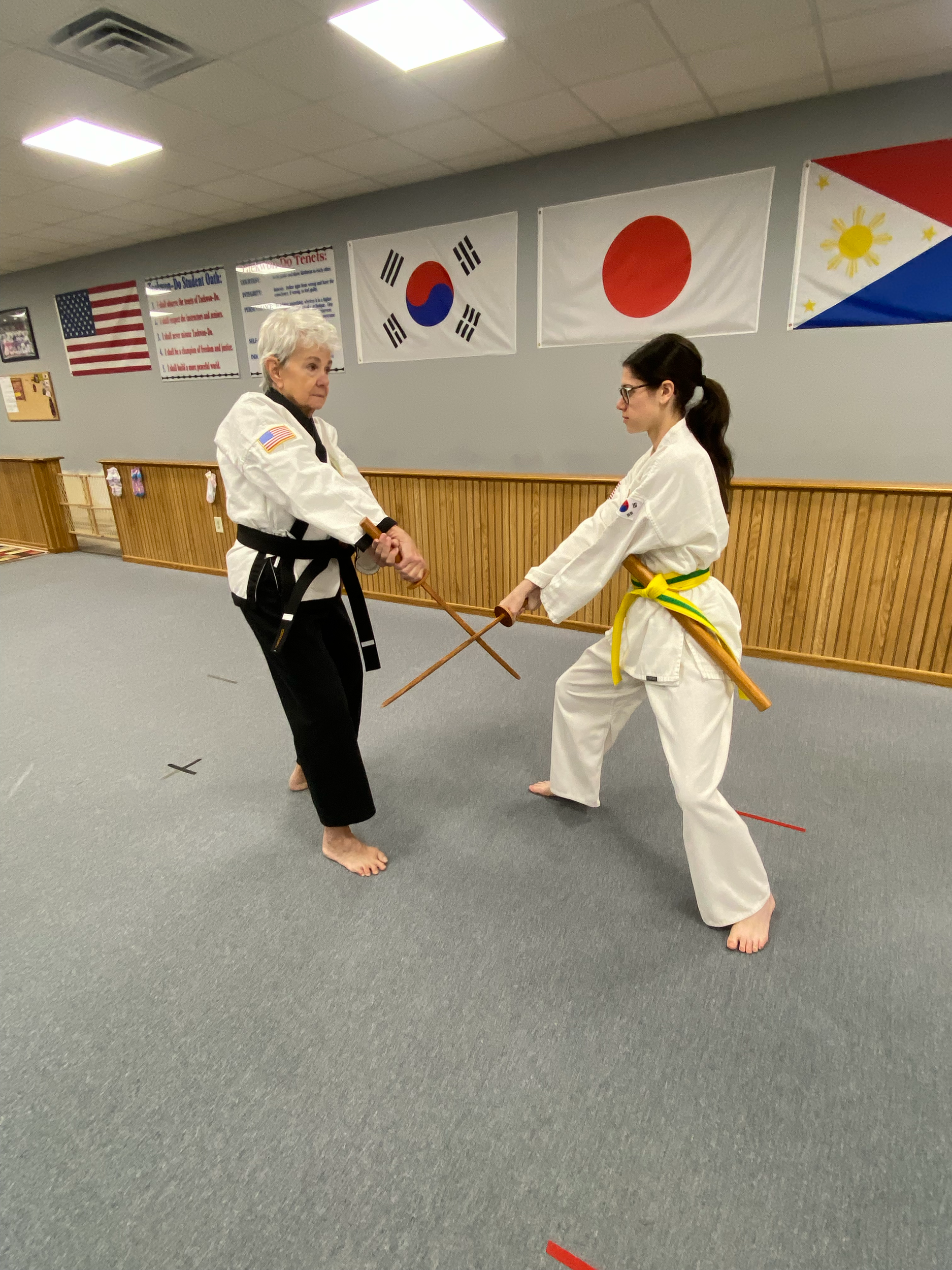 Two martial artists practicing martial arts in a dojo with flags of Japan, South Korea, and the United States hanging on the wall.
