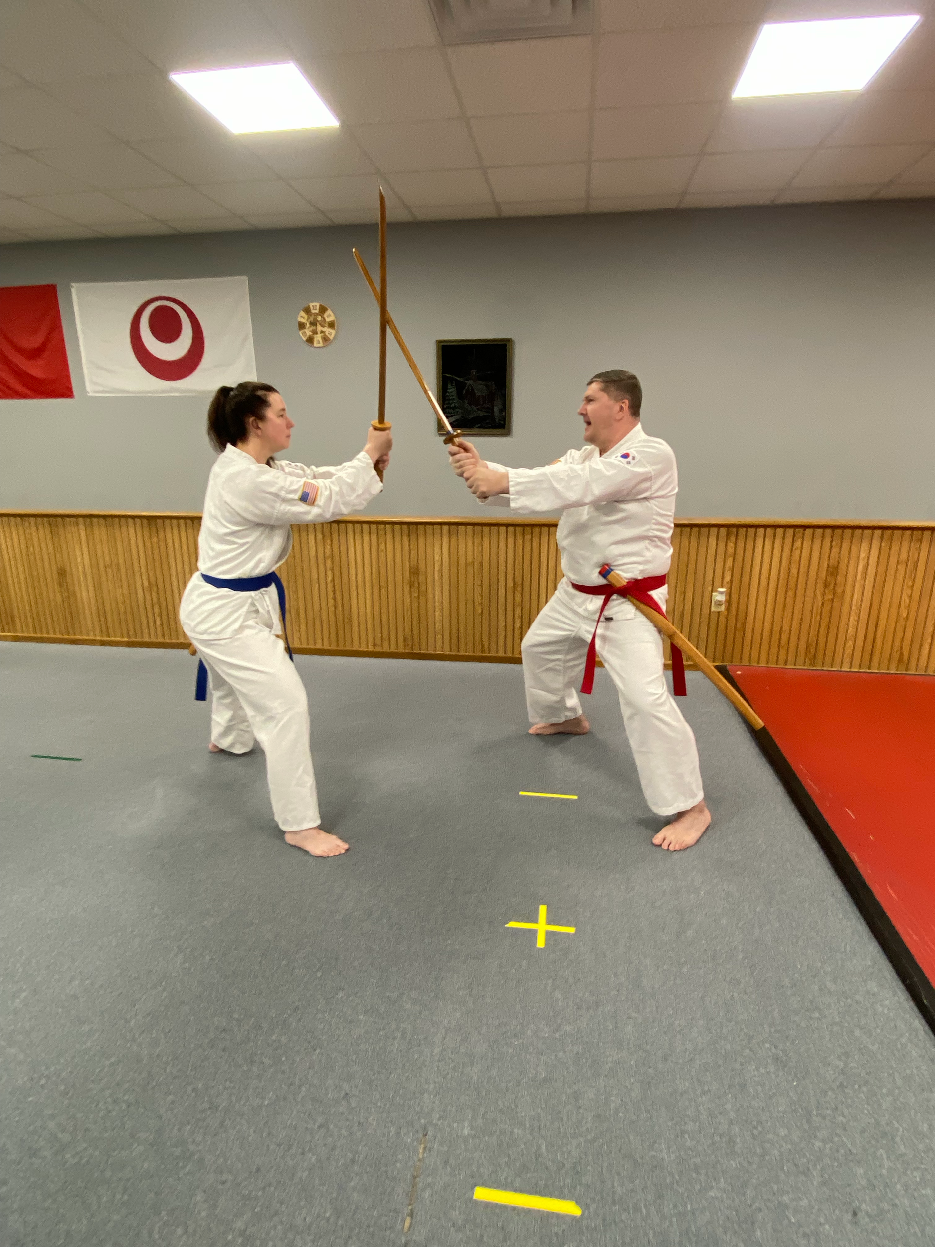 Two martial artists practicing a martial arts technique with wooden staffs in a dojo. One is a male with a red belt and the other a female with a blue belt, both wearing traditional white gis. They are standing on a gray mat with colored lines, in fr