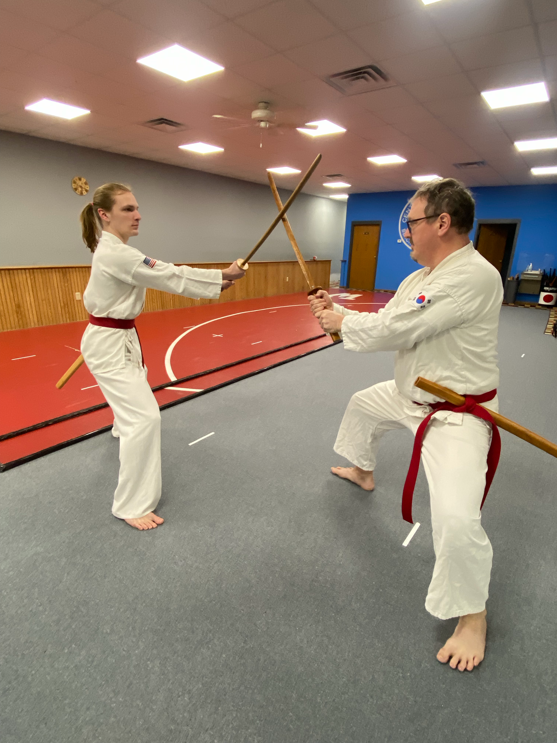Two martial artists in white uniforms practicing martial arts with wooden staffs in a gymnasium.