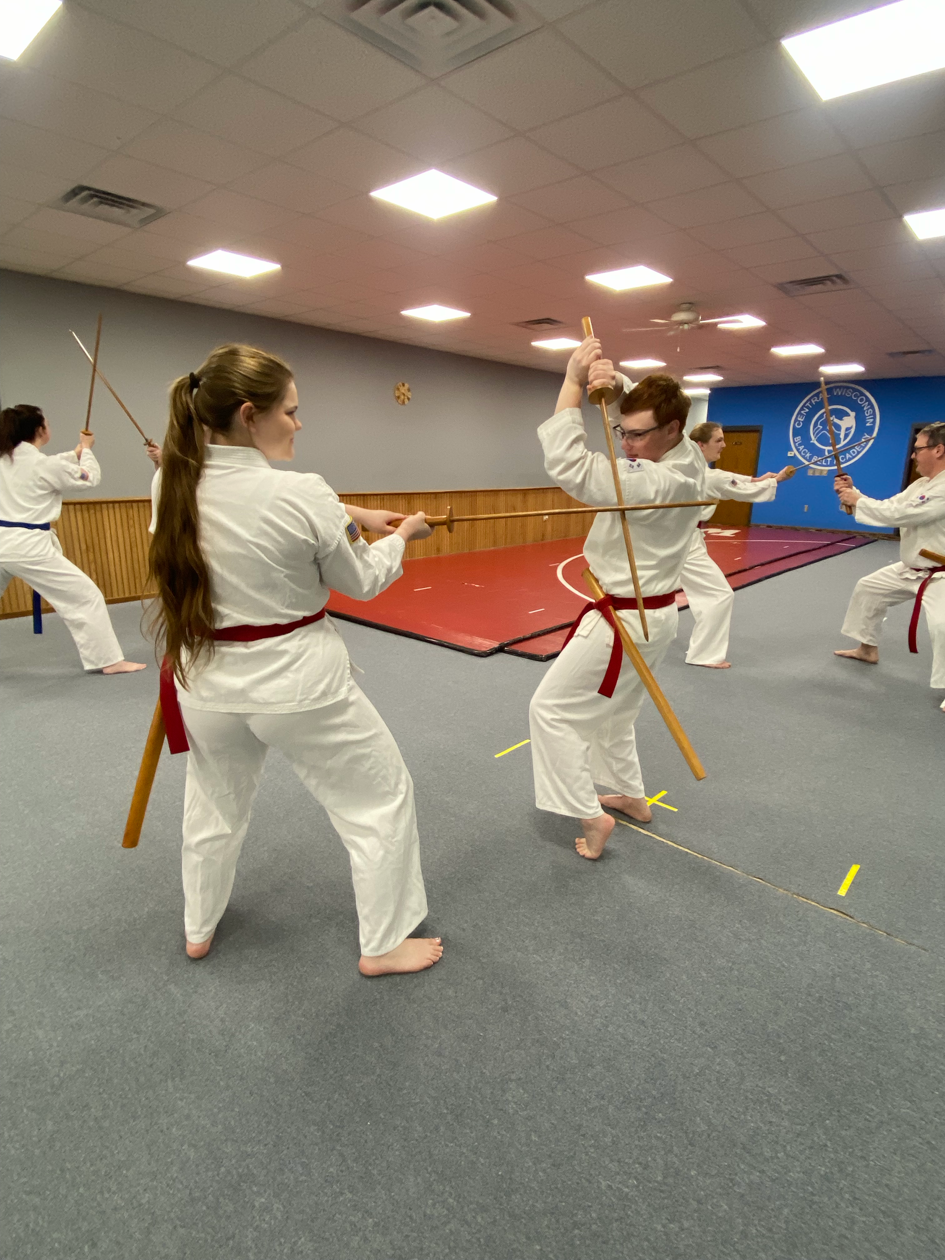 Two martial artists practicing kata with wooden staffs in a dojo, surrounded by others in white uniforms. The dojo has gray carpet flooring, a red and blue mat area, and a blue banner with a logo visible on the wall.