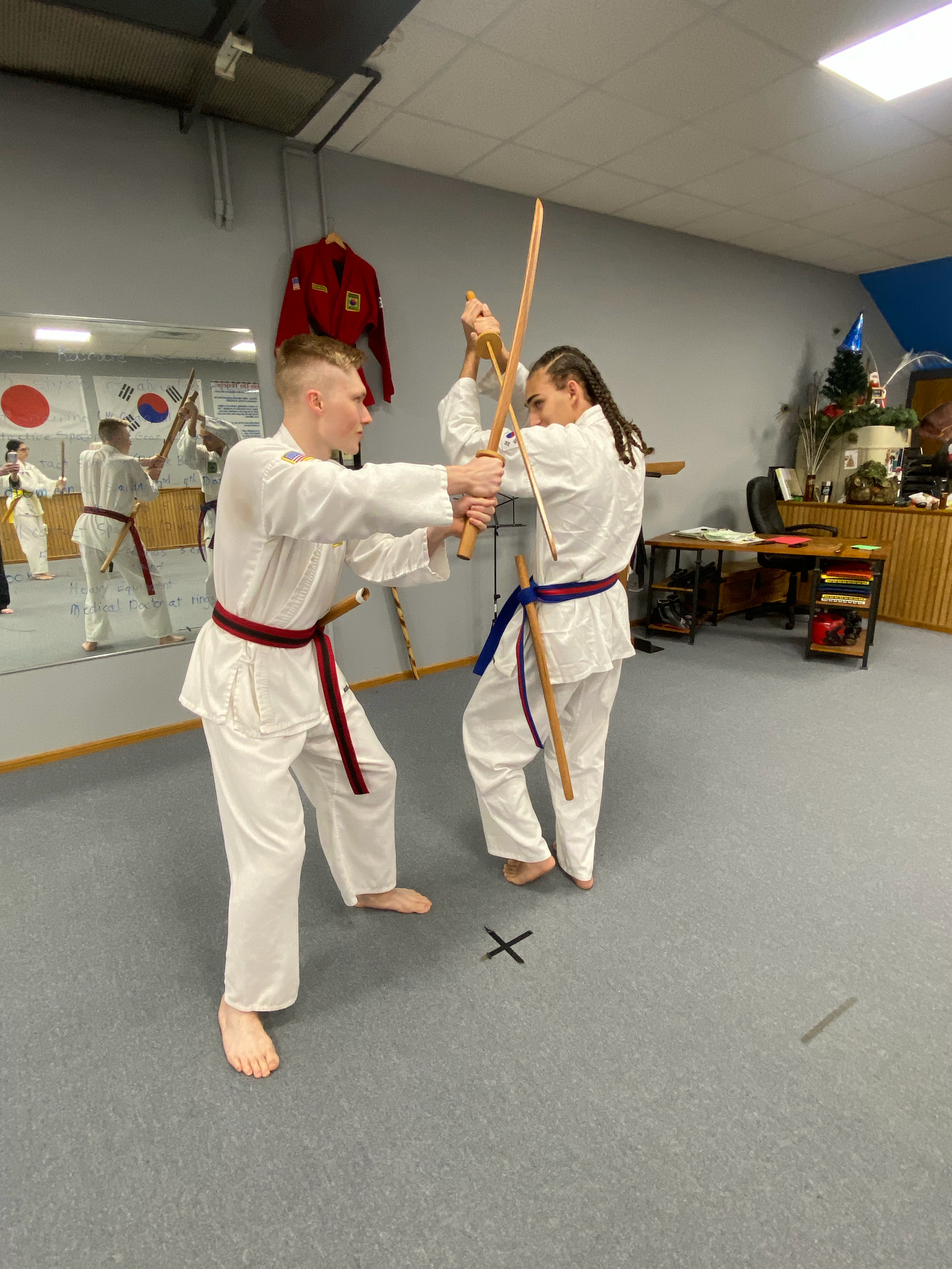 Two children practicing martial arts in a dojo, both wearing white gis with belts, one with a black belt, the other with a red belt. One child is holding a wooden staff, demonstrating a move, while the other watches. The room has gray flooring, a mir
