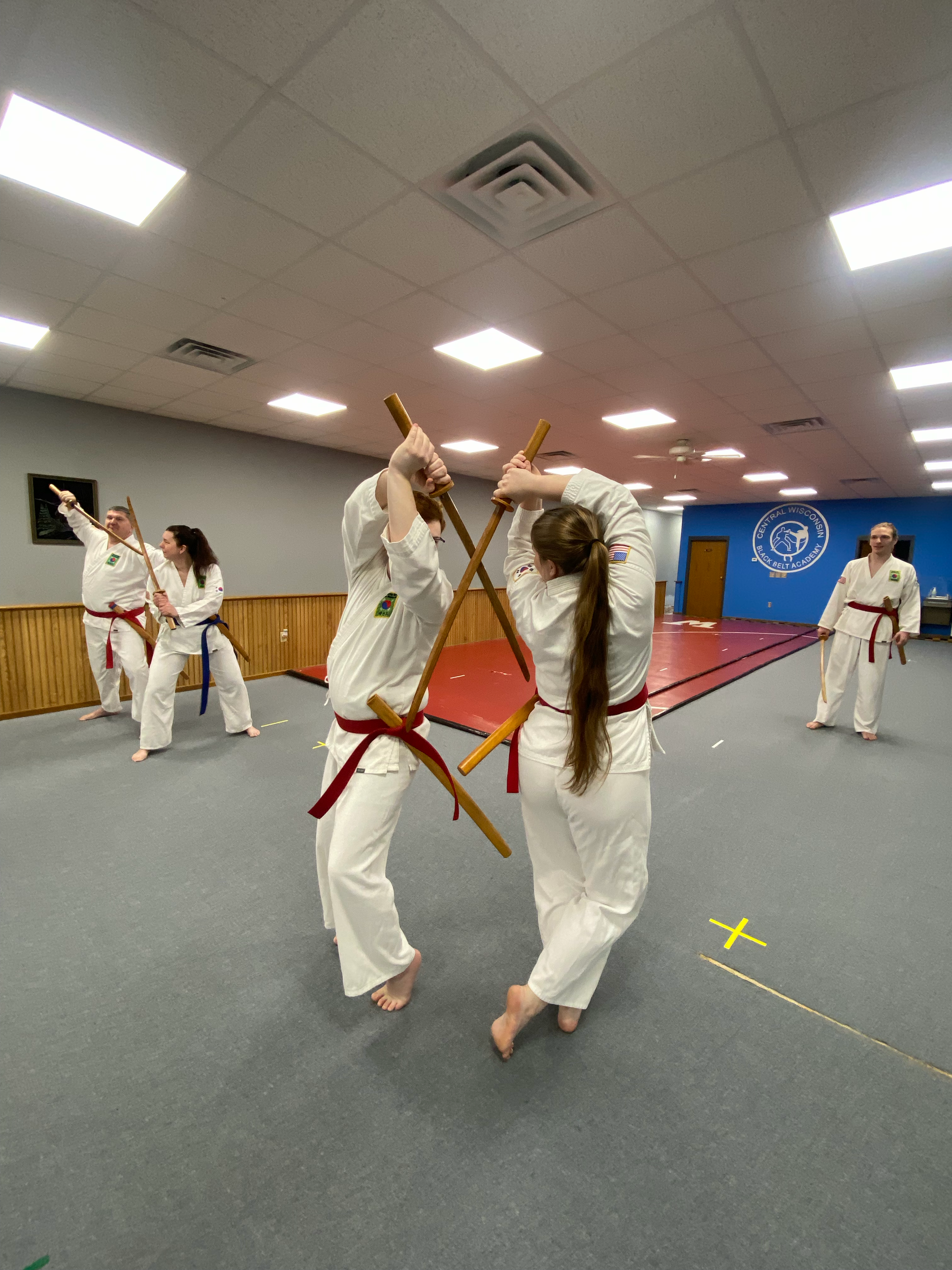 Martial arts students practicing with wooden sticks in a dojo, wearing white uniforms with red and blue belts.