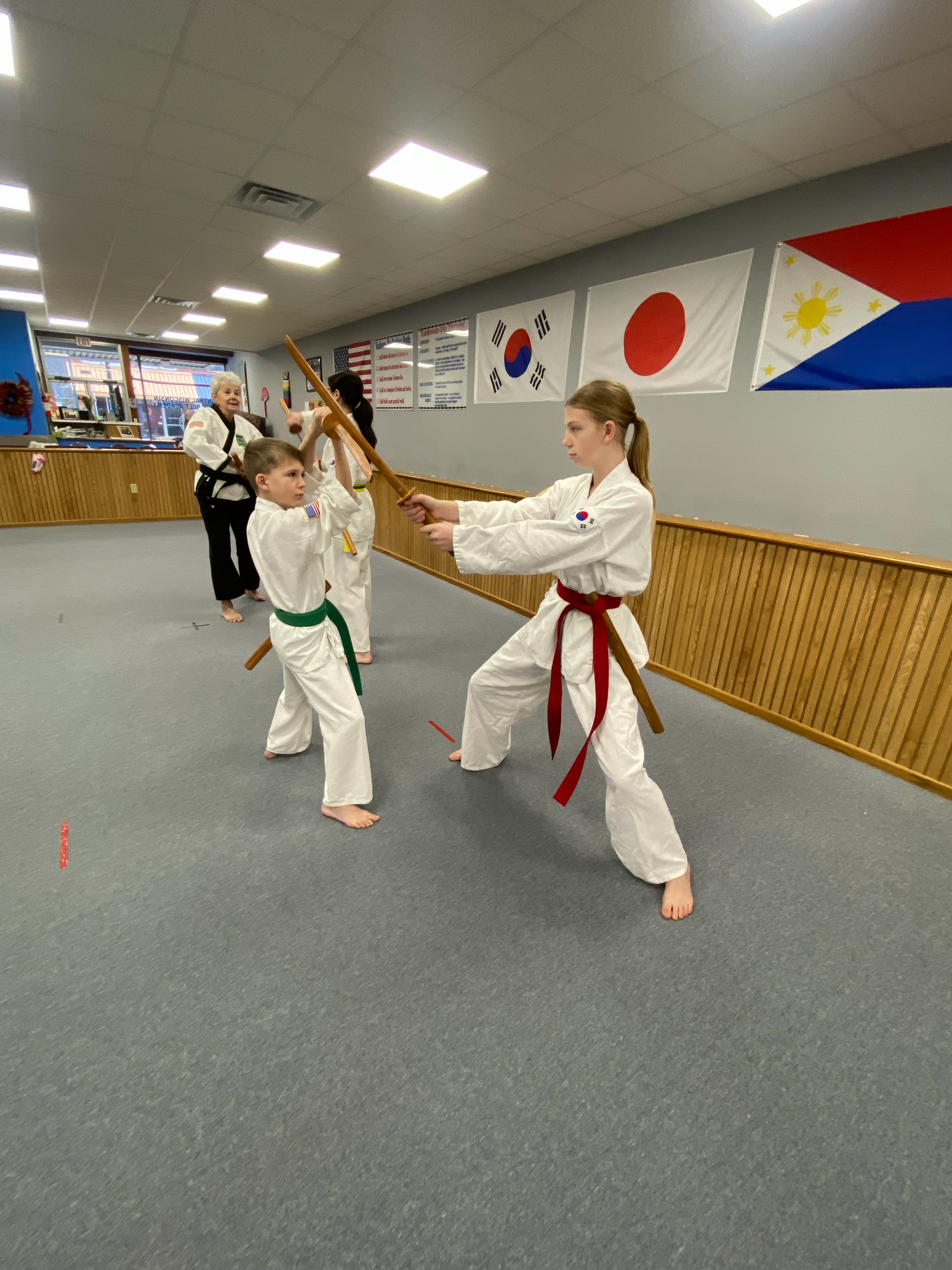 Two young girls practicing martial arts in a dojo, wearing traditional uniforms with red and green belts, holding wooden staffs, with flags of Japan, South Korea, the Philippines, and the United States hanging on the wall behind them.