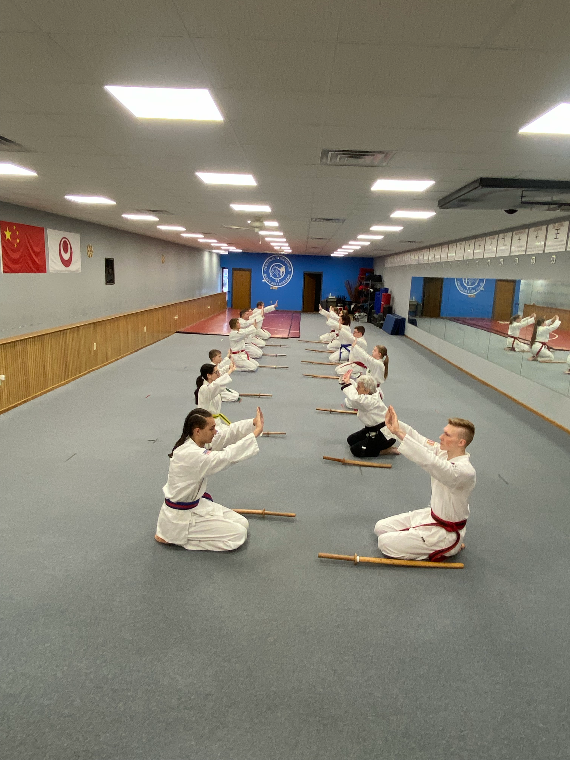 Martial arts students practicing in a dojo, sitting in seiza position on the floor, wearing traditional white uniforms with colored belts, facing their instructor who is demonstrating a move.