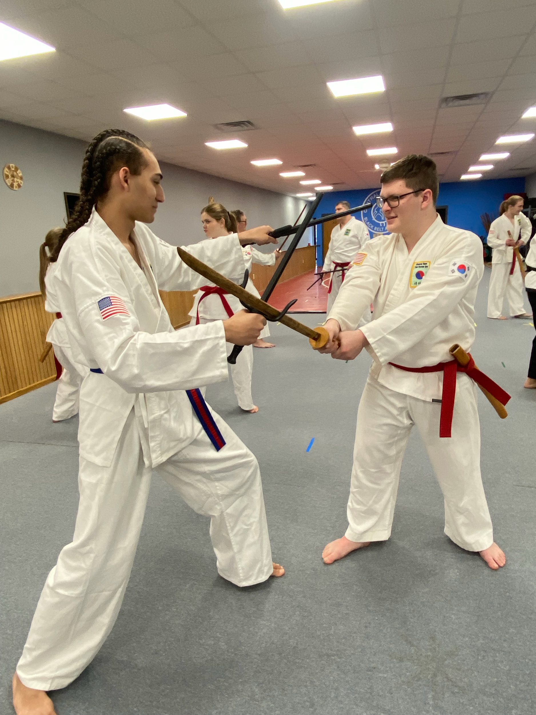 Martial arts students in white uniforms practicing with wooden sticks, inside a dojo with blue and brown walls and a gray floor. One student with glasses and braided hair demonstrates a technique while others observe.