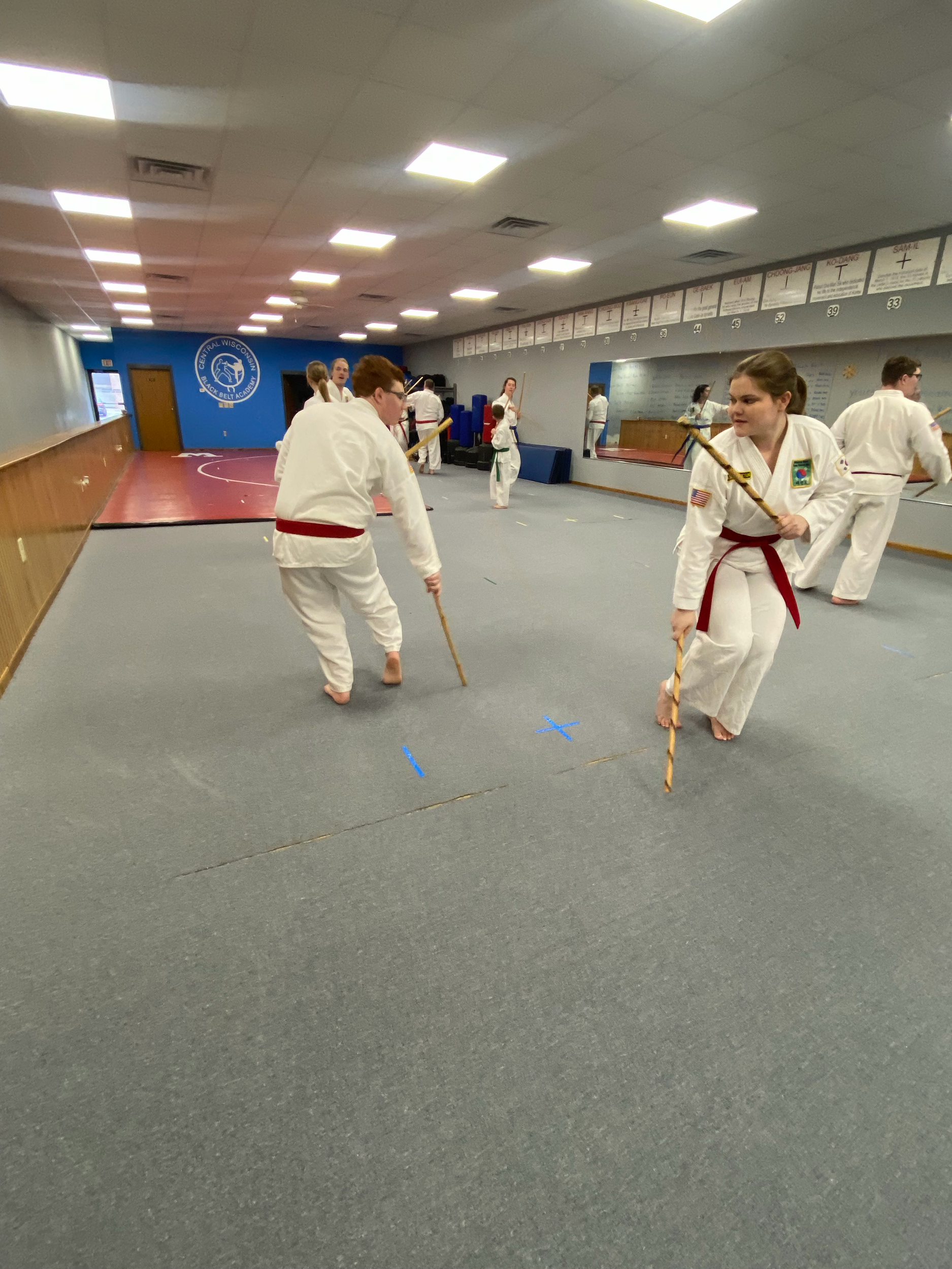 Children practicing martial arts in a dojo, wearing white uniforms with red belts, holding wooden sticks, with a blue emblem on the wall in the background.