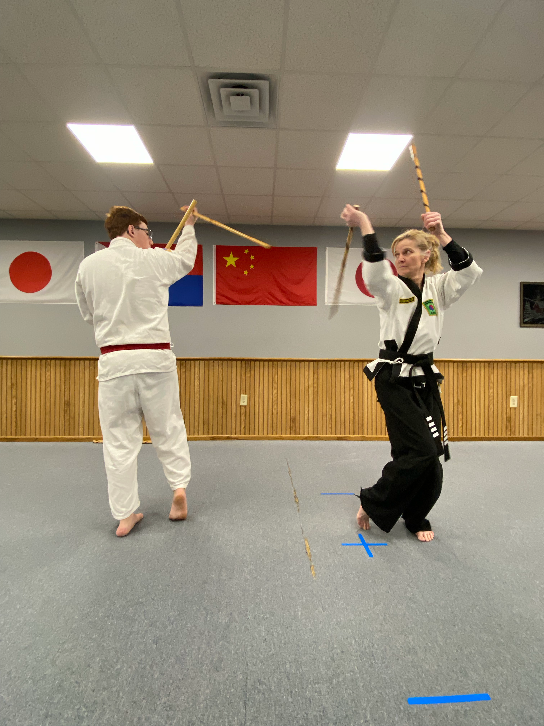 Two martial artists practicing with wooden sticks in a martial arts dojo. One is wearing a black uniform and the other is in a white uniform. Japanese, Chinese, and Brazilian flags are displayed in the background.
