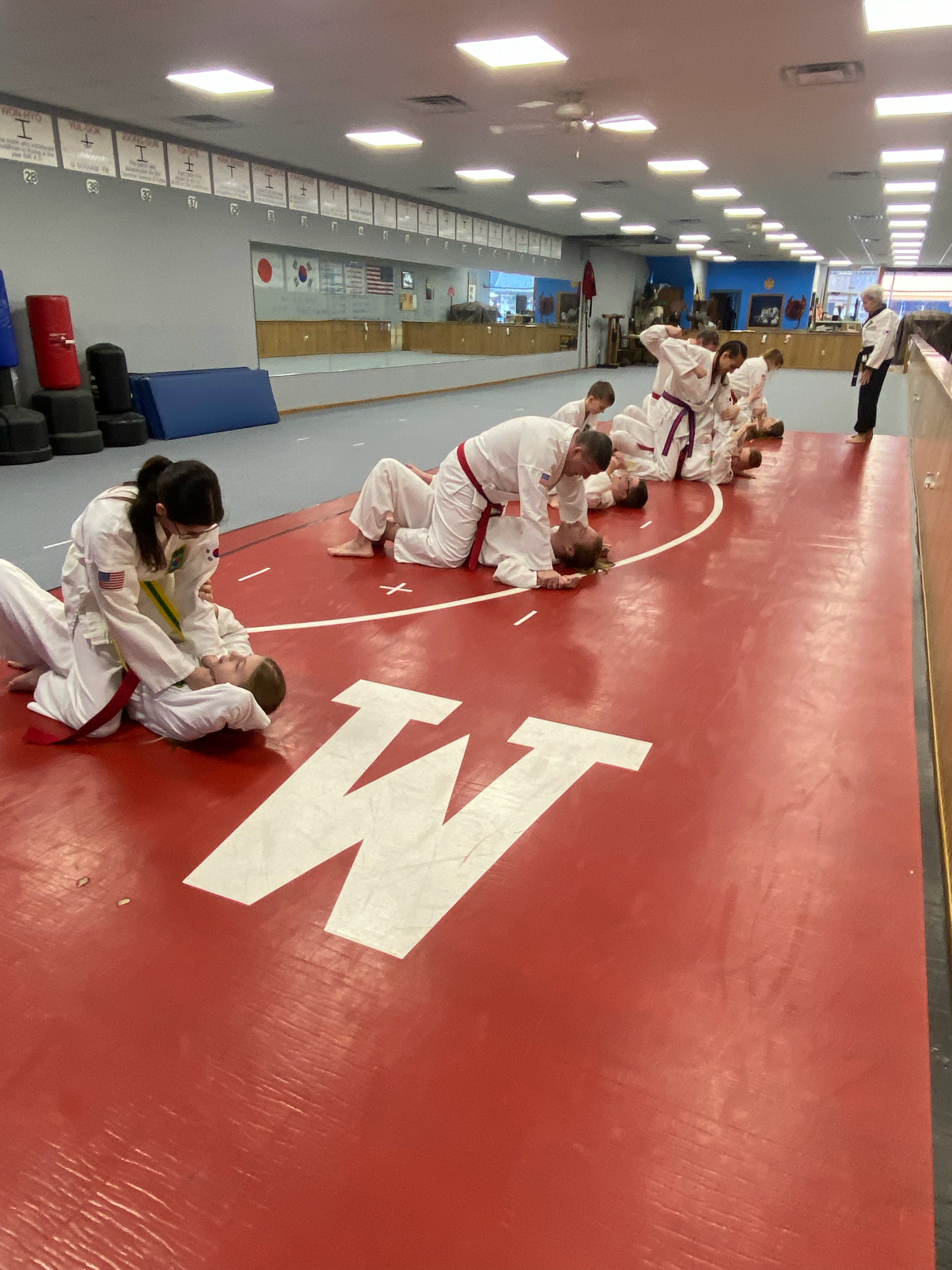 Kids practicing martial arts in a dojo, some sitting on the mat and others standing, with instructors and training equipment visible.