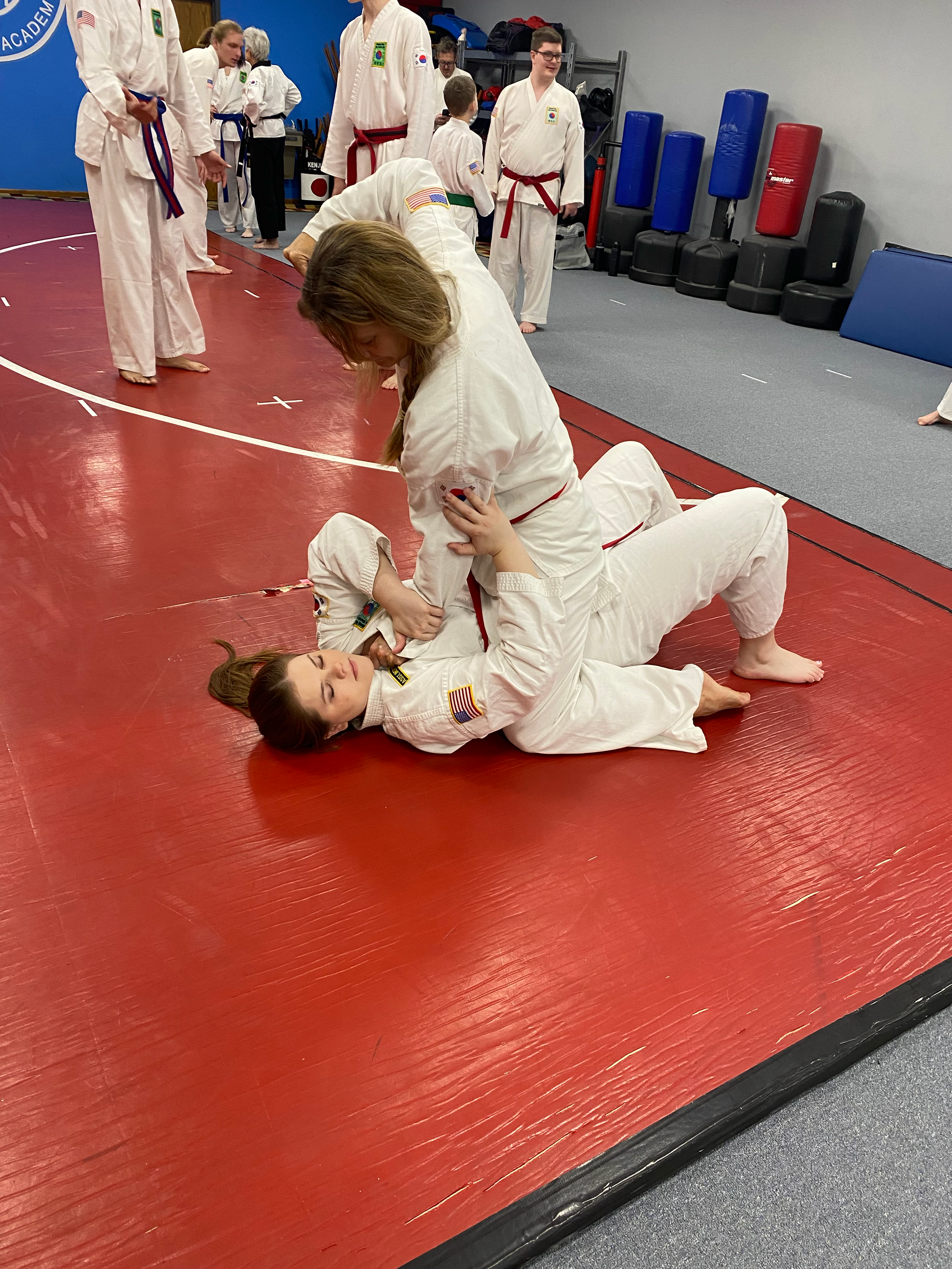 Two women practicing Brazilian Jiu-Jitsu on a red mat in a martial arts gym. The woman on the ground is in a white gi, while the woman standing is in a white gi with red trim. Other practitioners and training equipment are visible in the background.