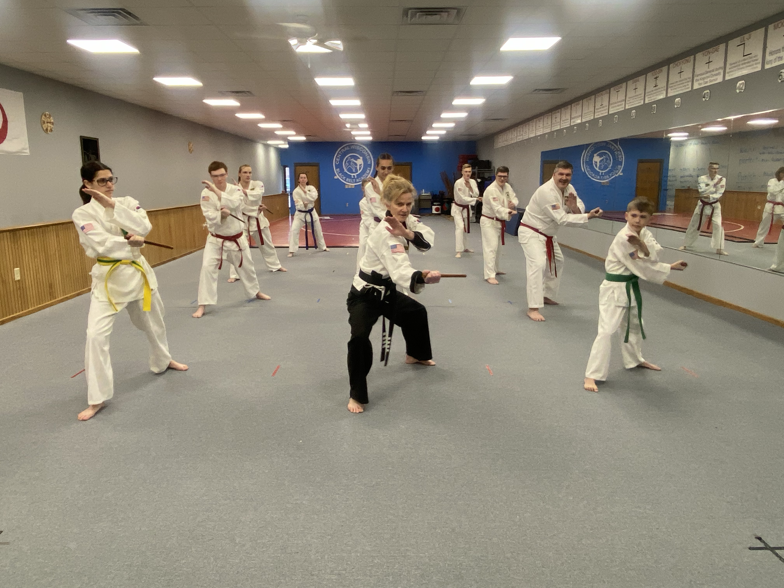 Group of martial arts students practicing in a dojo, wearing white uniforms with colored belts, with an instructor demonstrating with a wooden sword at the front. The dojo has gray walls, a mirrored wall, and a blue logo on the back wall.