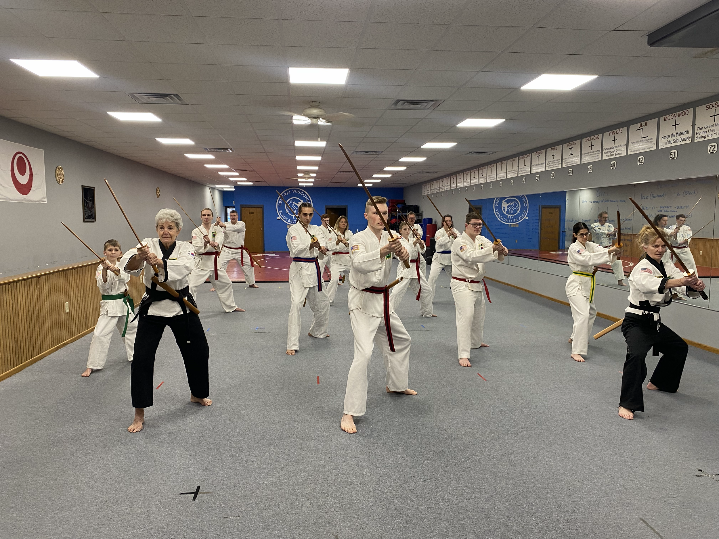 Group of people practicing martial arts with wooden staffs in a dojo, including children and adults, wearing karate uniforms with colored belts.