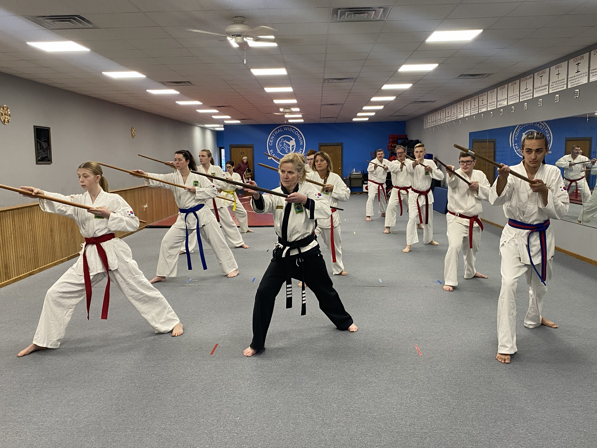 Group of martial arts students practicing with sticks in a dojo, guided by an instructor in black uniform at the front.